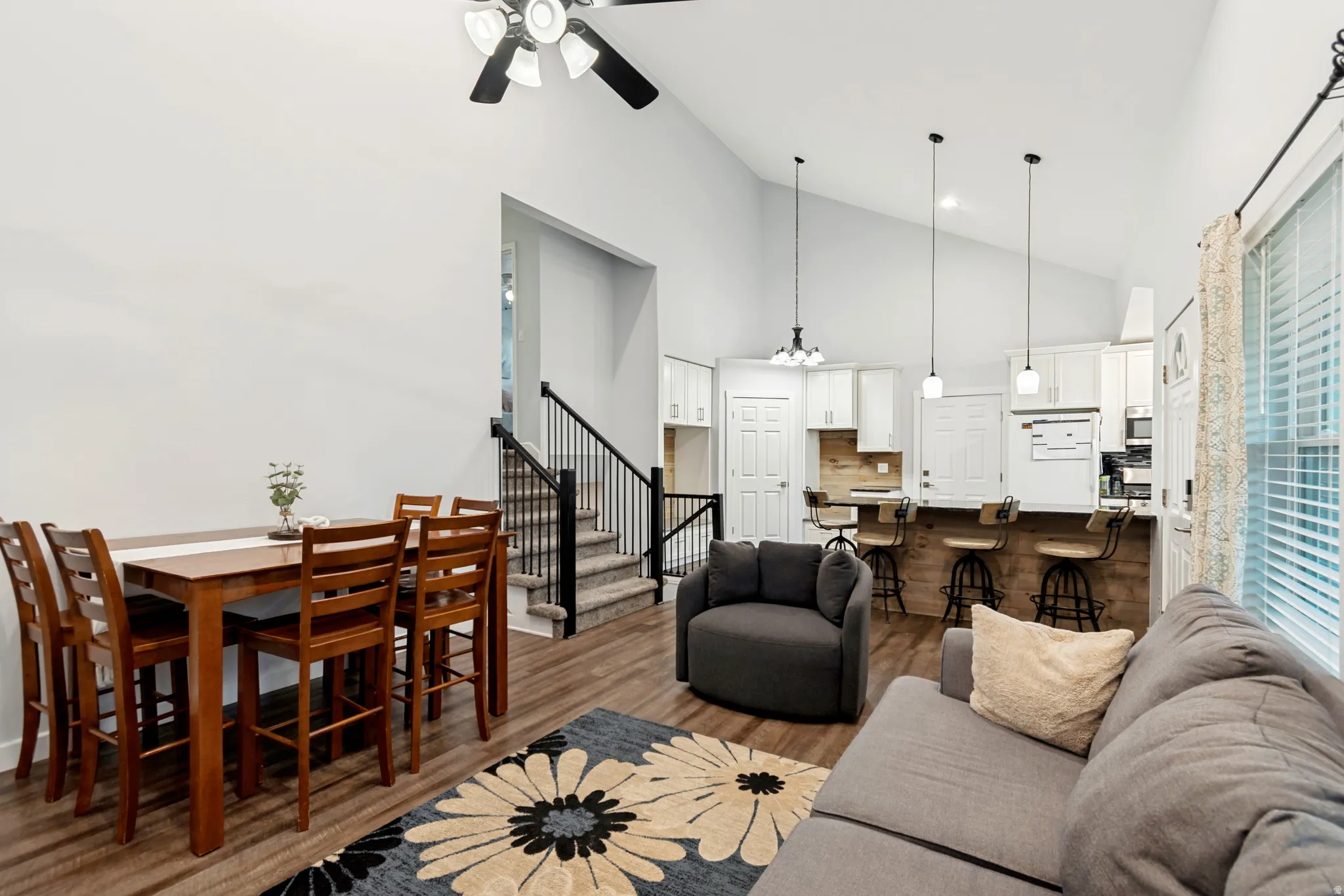 Open-concept living area featuring a vaulted ceiling with a ceiling fan, wood-finish flooring, a kitchen with white cabinetry and a breakfast bar, and a staircase with metal railings