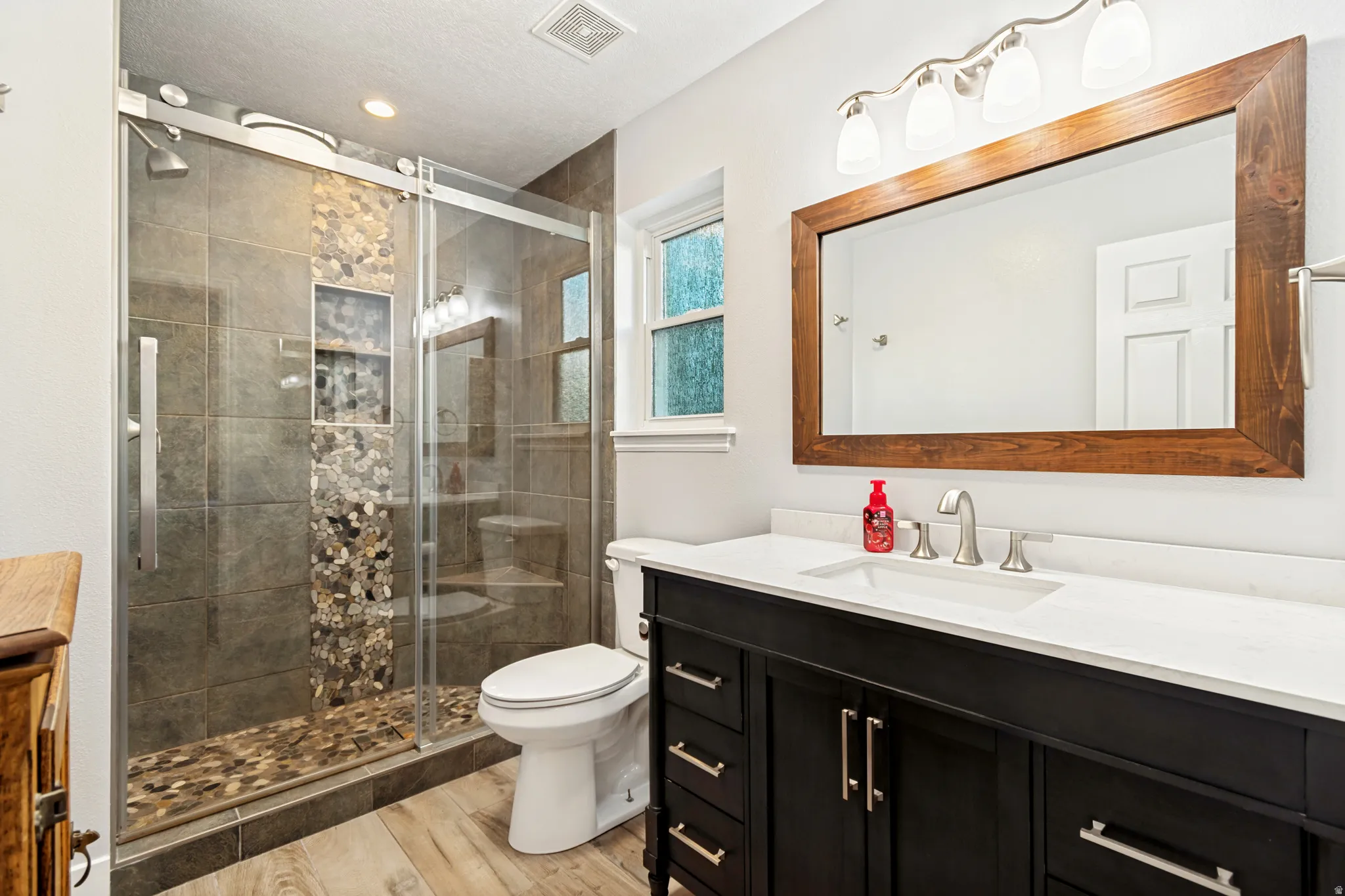 Spacious bathroom featuring a glass-enclosed shower with pebble-tiled floor and accent strip, a dark wood-finish vanity with white countertop and integrated sink, a large wood-framed mirror, and wood-finish flooring