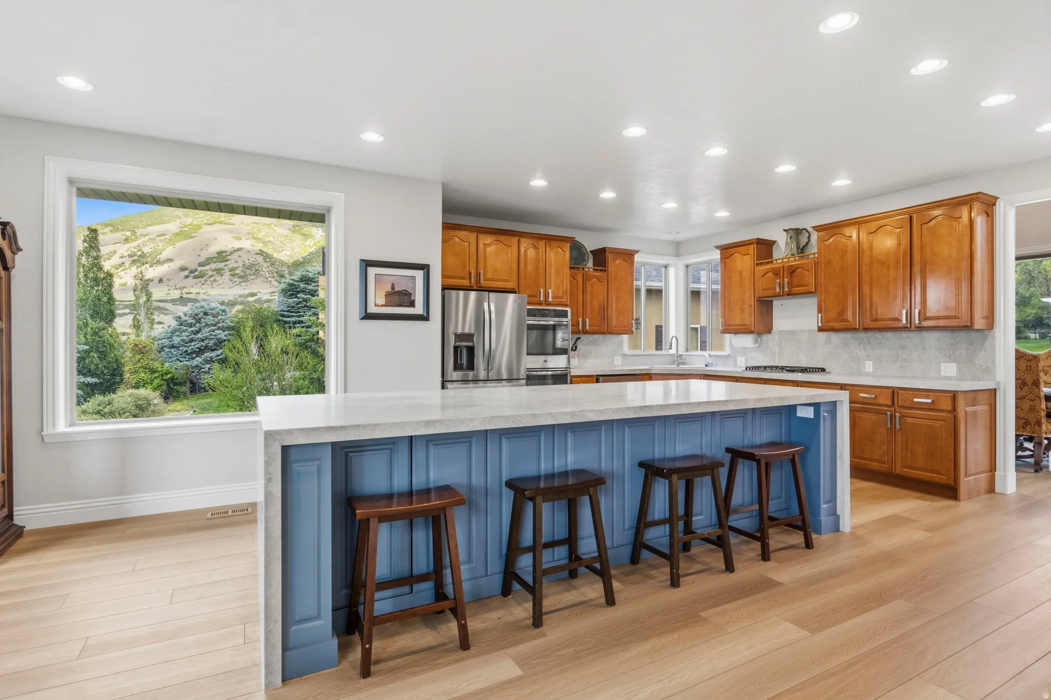 Expansive kitchen featuring a large central island with a light-toned countertop, wood-finish flooring, recessed lighting, wood cabinetry, and stainless steel appliances