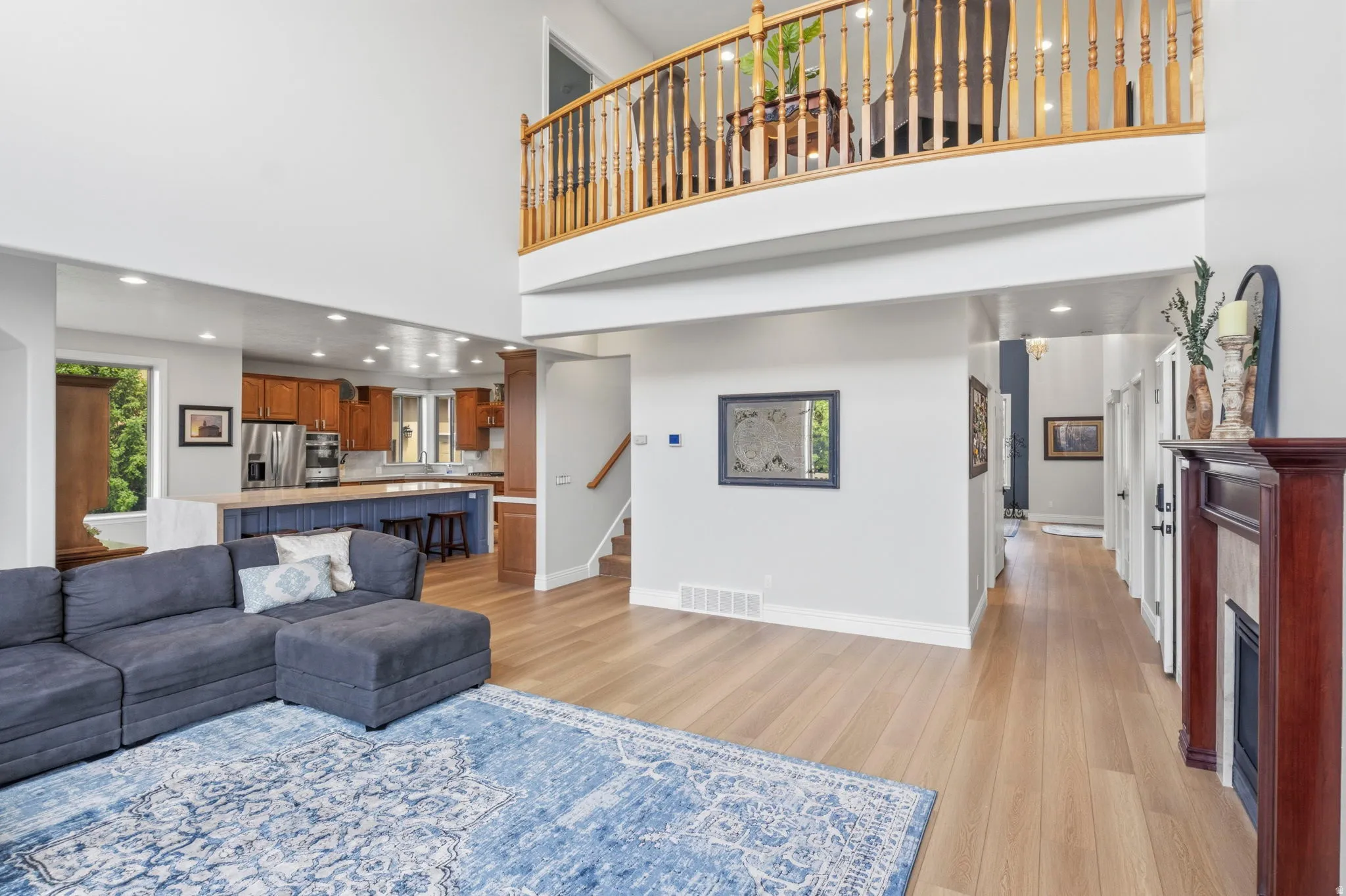 Open-concept living space featuring high ceilings, wood-finish flooring, a kitchen with wood cabinetry and a central island, a fireplace with a wood mantle, and a second-story interior balcony with wood railings