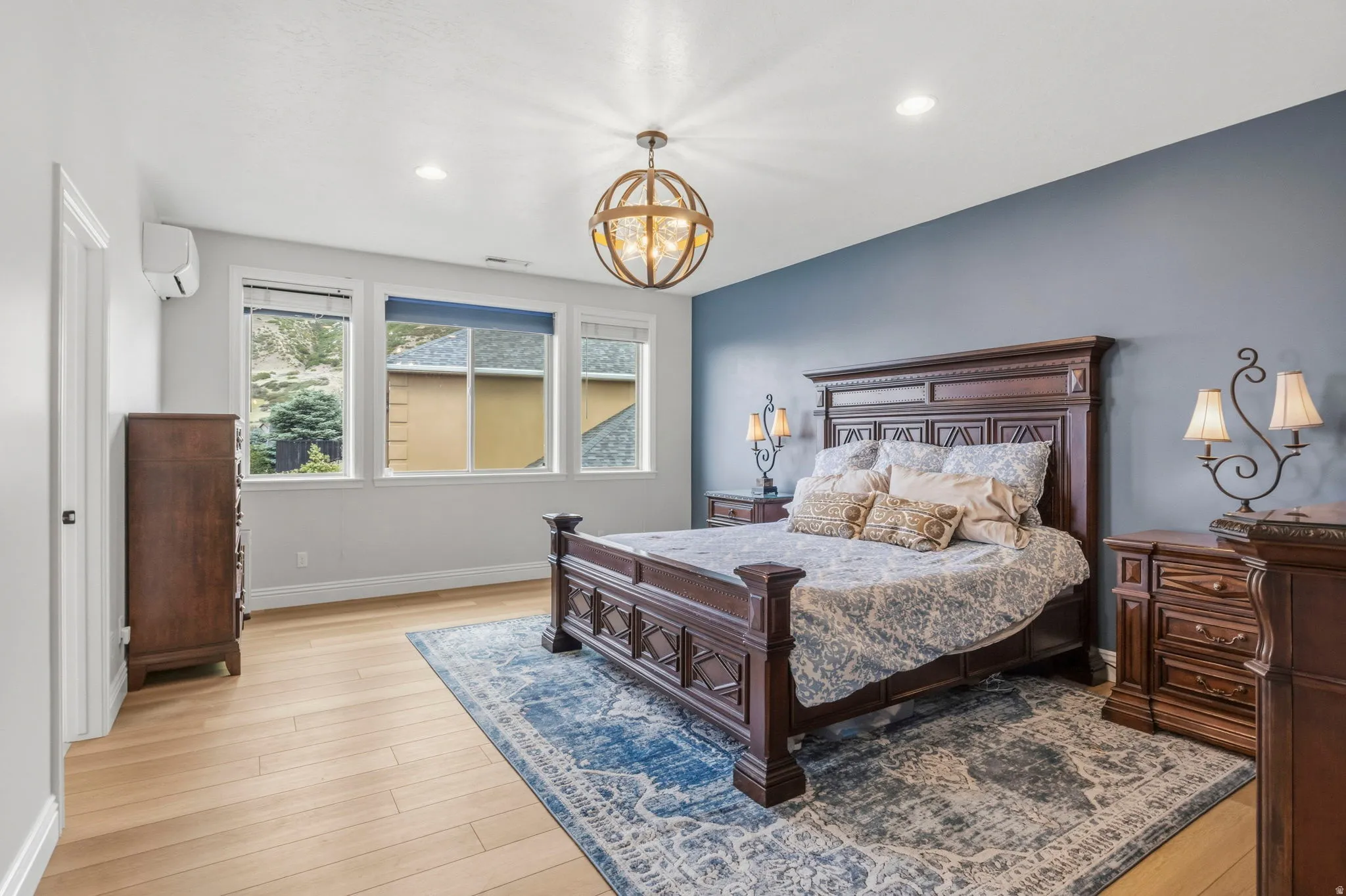 Primary Bedroom. Spacious room featuring wood-finish flooring, a contemporary ceiling light fixture, three large windows, and a built-in wall-mounted climate control unit