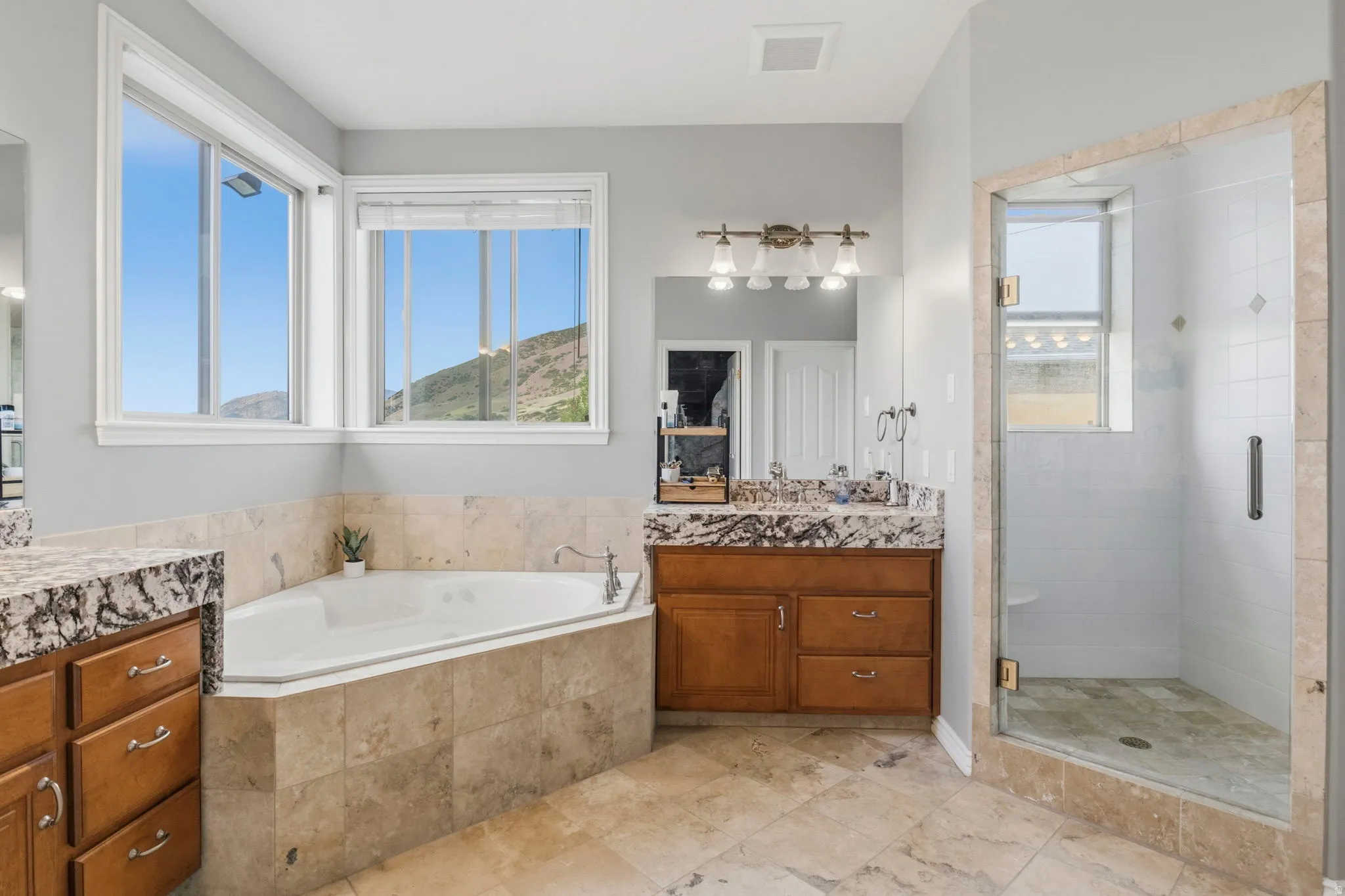 Spacious bathroom featuring a corner soaking tub with tile surround, a glass-enclosed shower, wood cabinetry with granite-finish countertops, and large format tile flooring