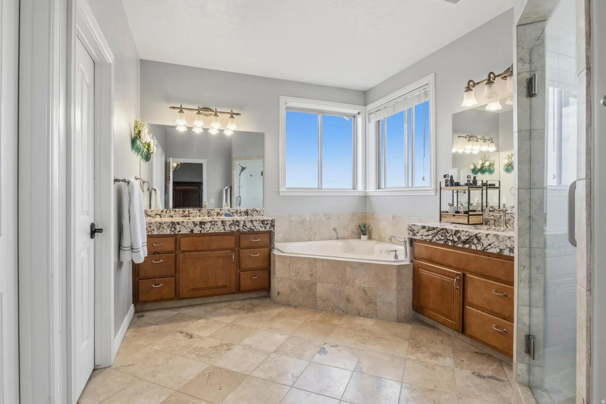 Primary Bathroom. Spacious bathroom featuring light-toned tile flooring, a corner soaking tub, two separate vanity areas with granite countertops and wood cabinetry, and a glass-enclosed shower