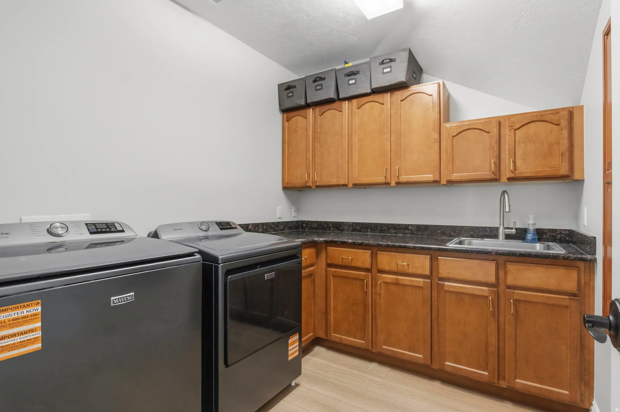 Dedicated laundry area featuring wood-finish flooring, ample cabinetry for storage, a stainless steel sink, and dark countertops