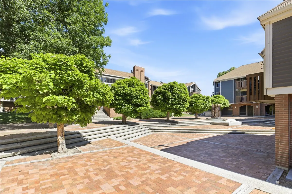 Expansive brick paver courtyard featuring mature trees, terraced concrete steps, and a multi-level building with brick and blue siding accents