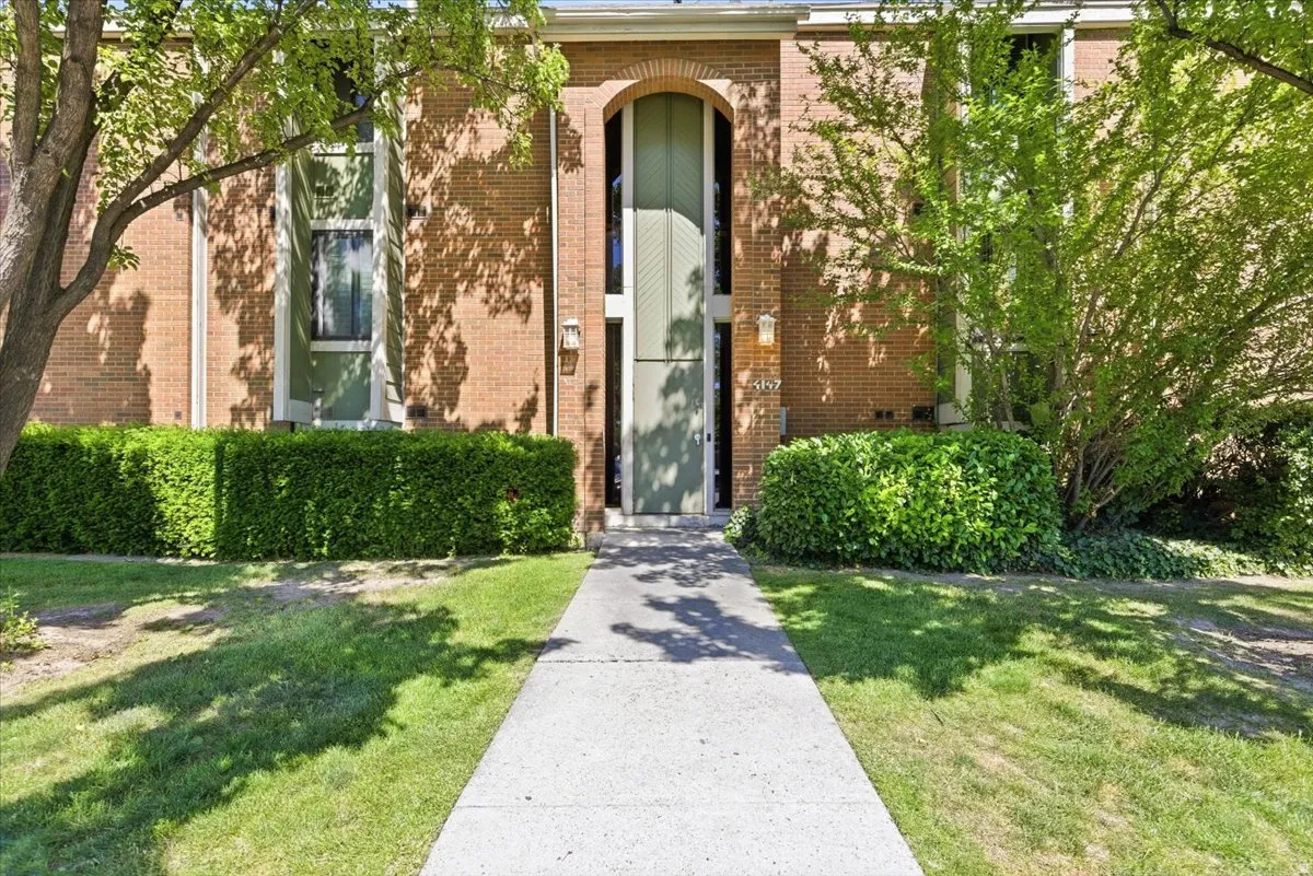 Brick exterior featuring an arched entryway, established landscaping, a concrete pathway, and mature shade trees