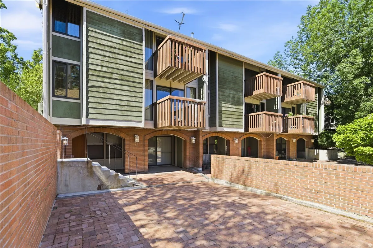 Multi-level building with brick and horizontal siding facade, featuring a paver driveway, multiple wooden balconies, and arched ground-level entryways
