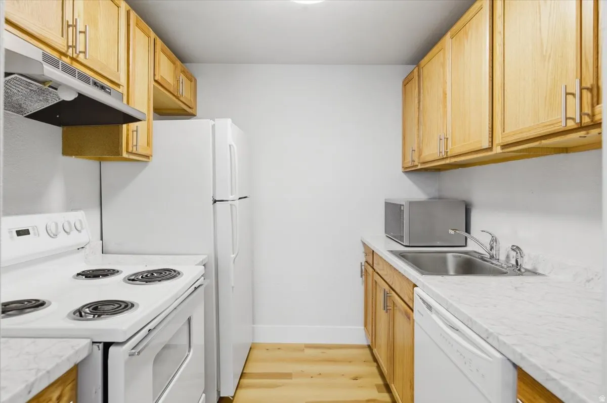 Galley kitchen featuring light wood cabinetry, white appliances, durable countertops, and wood-finish flooring