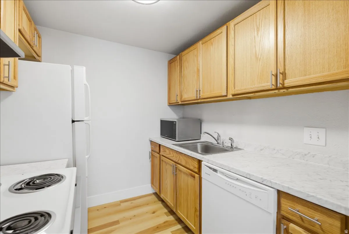 Compact kitchen featuring natural wood cabinetry, light-toned countertops, a stainless steel sink, a white dishwasher, and wood-finish flooring