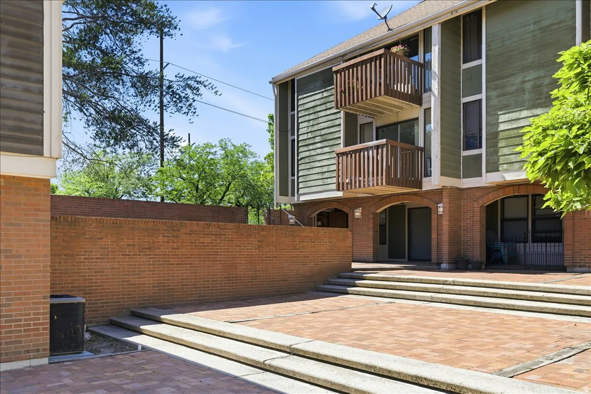 Multi-level building exterior featuring brick and green siding, ground-level arched entrances, upper-level balconies with wood railings, and a brick paver patio with concrete steps