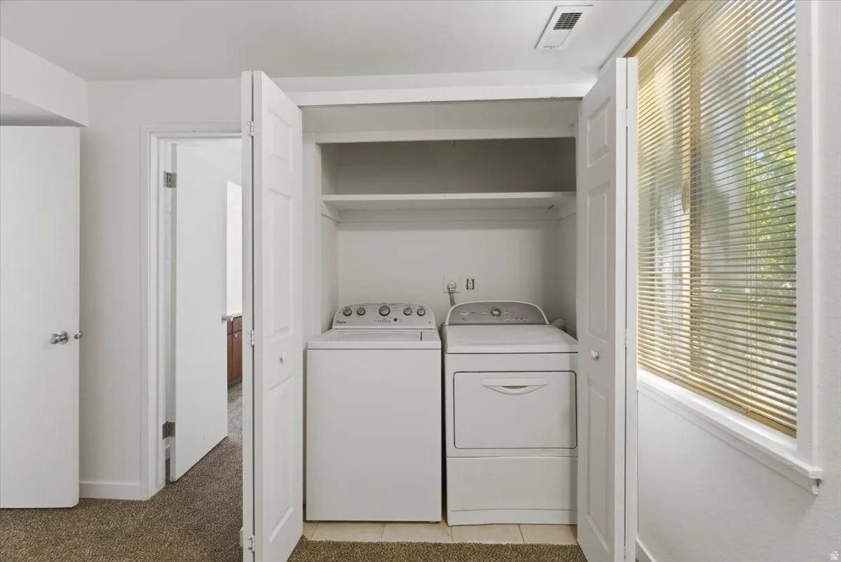In-unit laundry closet featuring a stacked washer and dryer, upper shelving, tiled flooring, and nearby natural light from a window with blinds