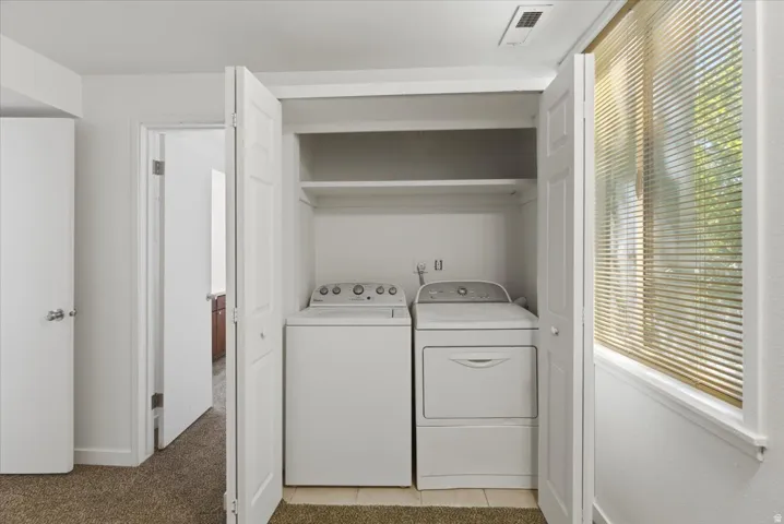 In-unit laundry closet featuring a stacked washer and dryer, upper shelving, tiled flooring, and nearby natural light from a window with blinds