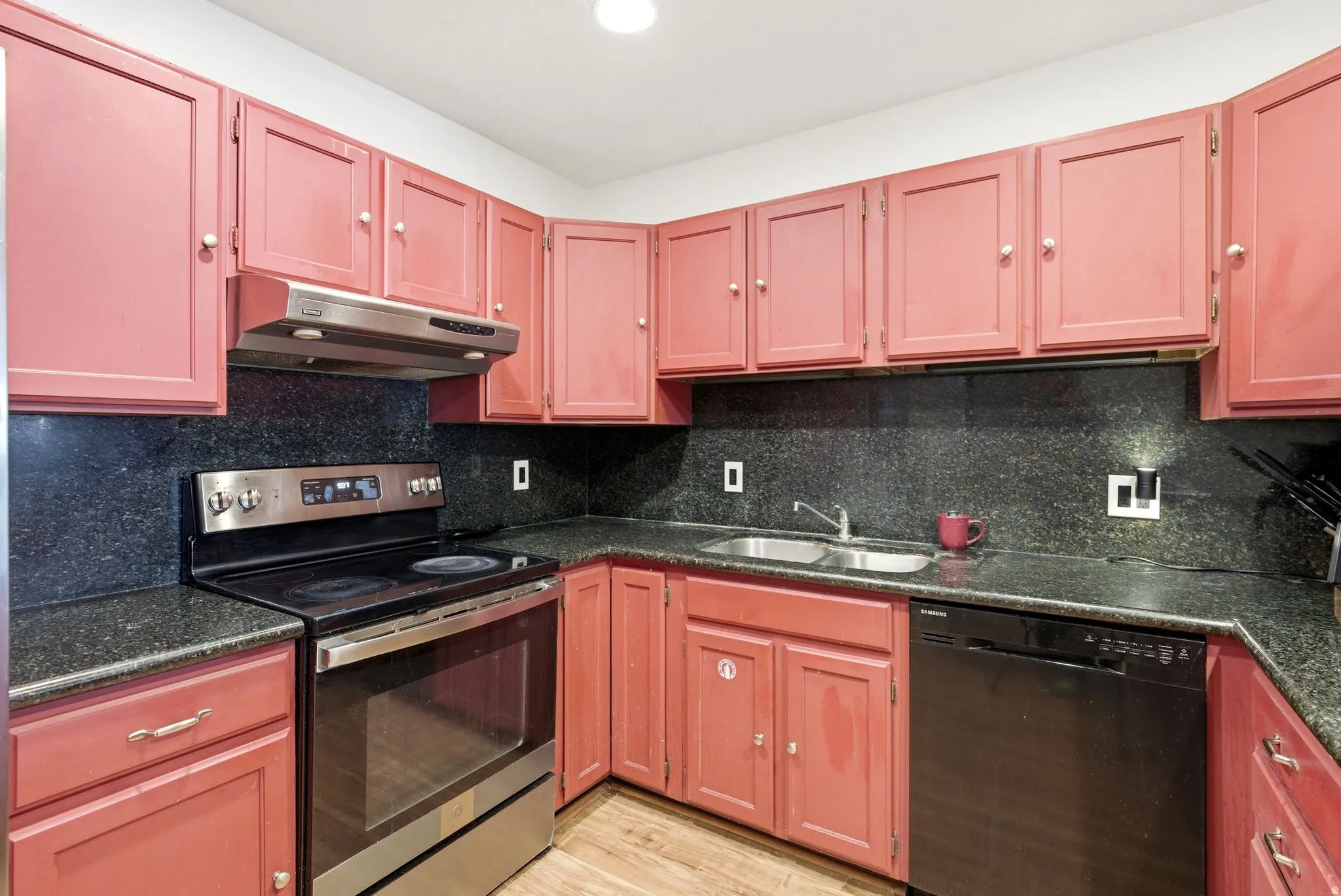 Kitchen featuring red cabinetry, dark stone countertops, stainless steel range, coordinating range hood, and a dual basin sink
