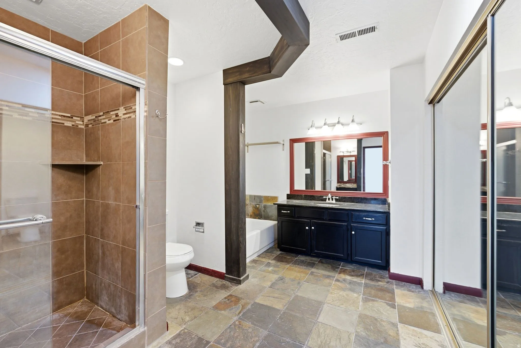 Spacious bathroom featuring a glass-enclosed shower with tile surround and integrated shelf, a vanity with dark cabinetry and countertop, a large framed mirror, and slate-finish floor tiles