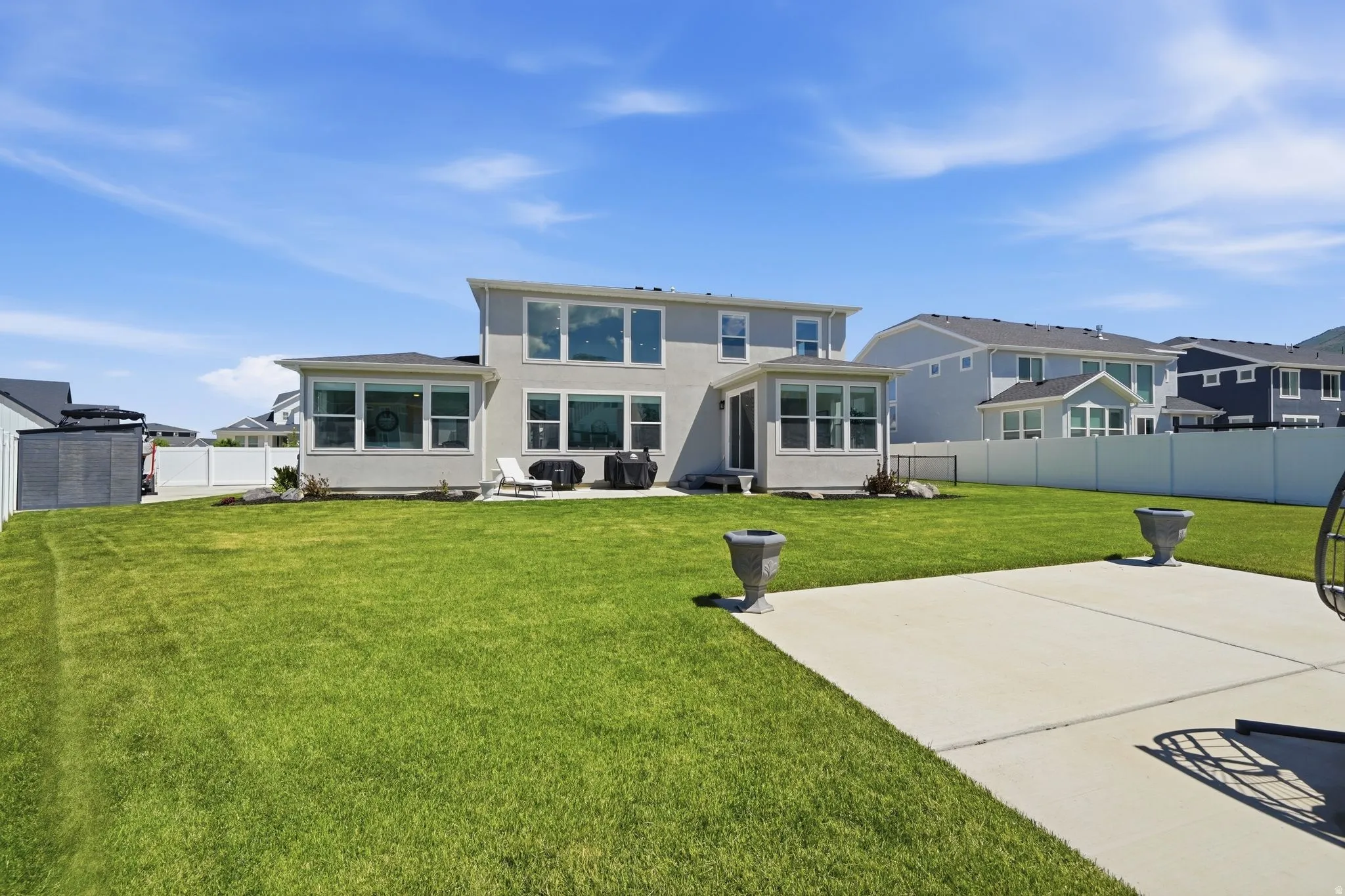 Expansive rear yard featuring a large concrete patio, manicured lawn, and a light-colored stucco exterior with multiple windows