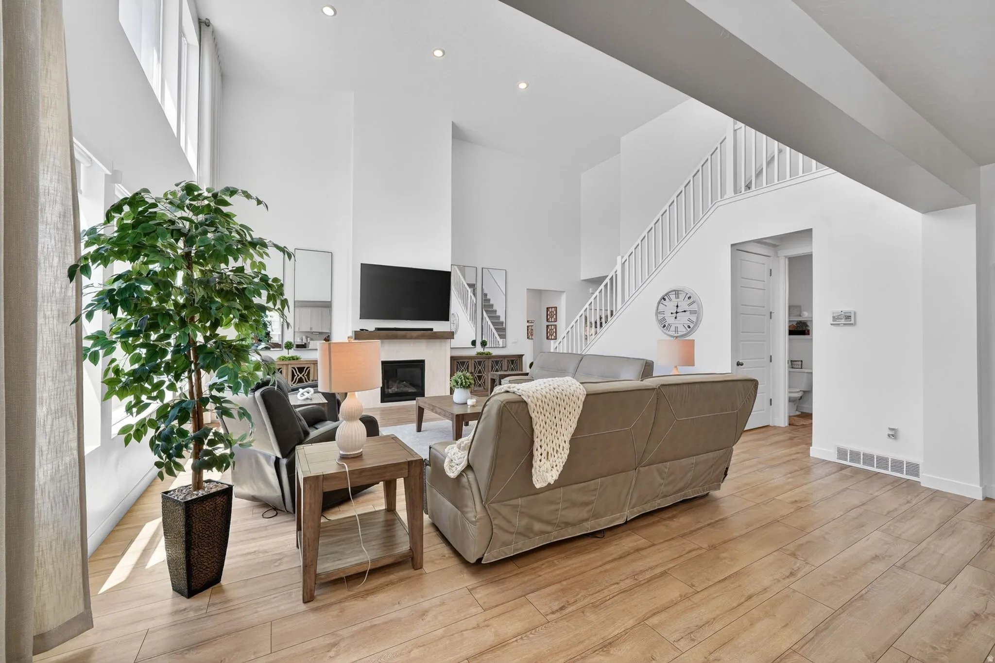 Open-concept living space featuring high ceilings, wood-finish flooring, a fireplace with a light-toned surround and wood mantel, a white staircase with balusters, and transom windows