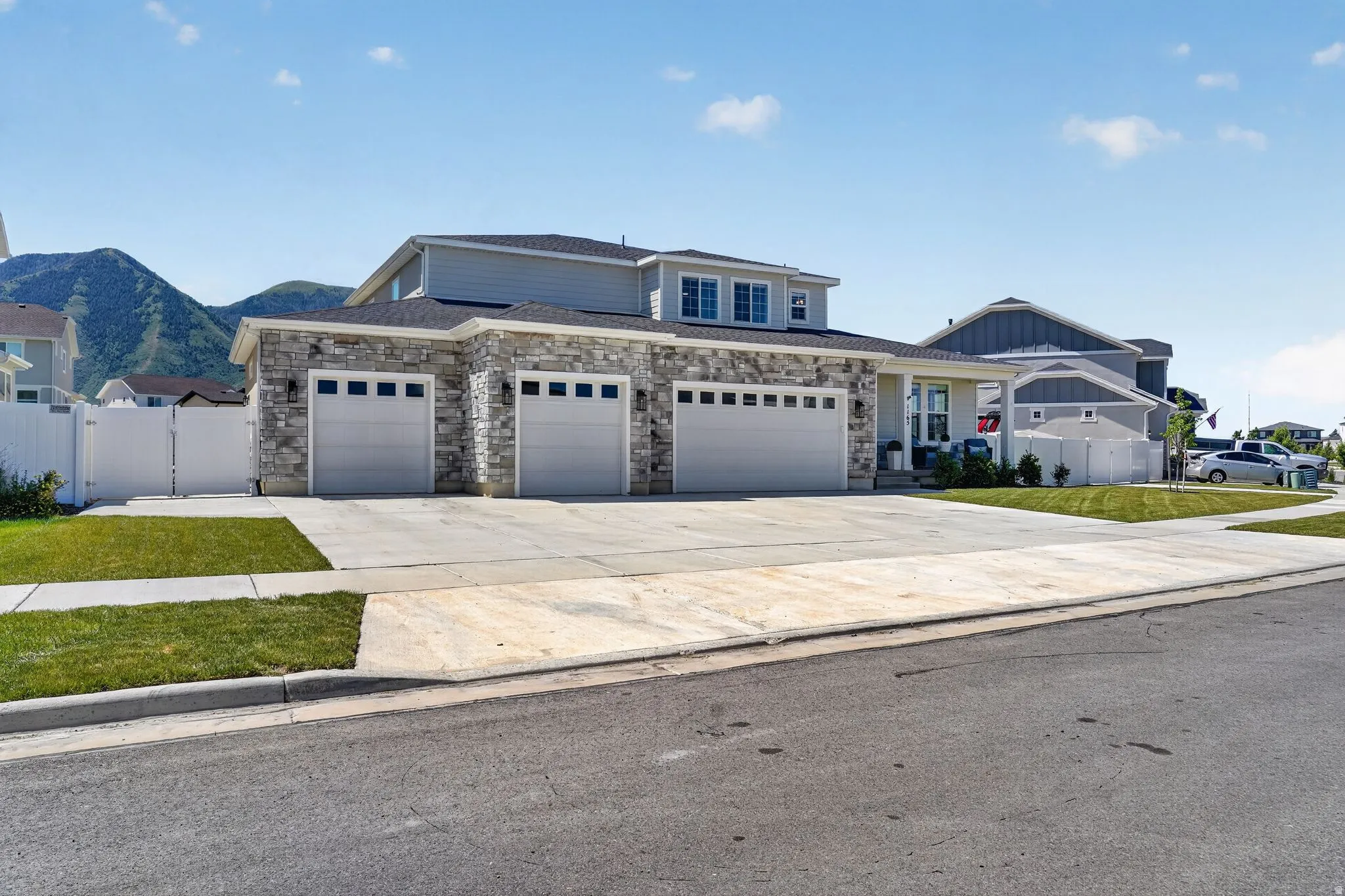 Modern multi-level residence featuring a gray siding and stacked stone facade with a three-car garage, concrete driveway, and white picket fencing