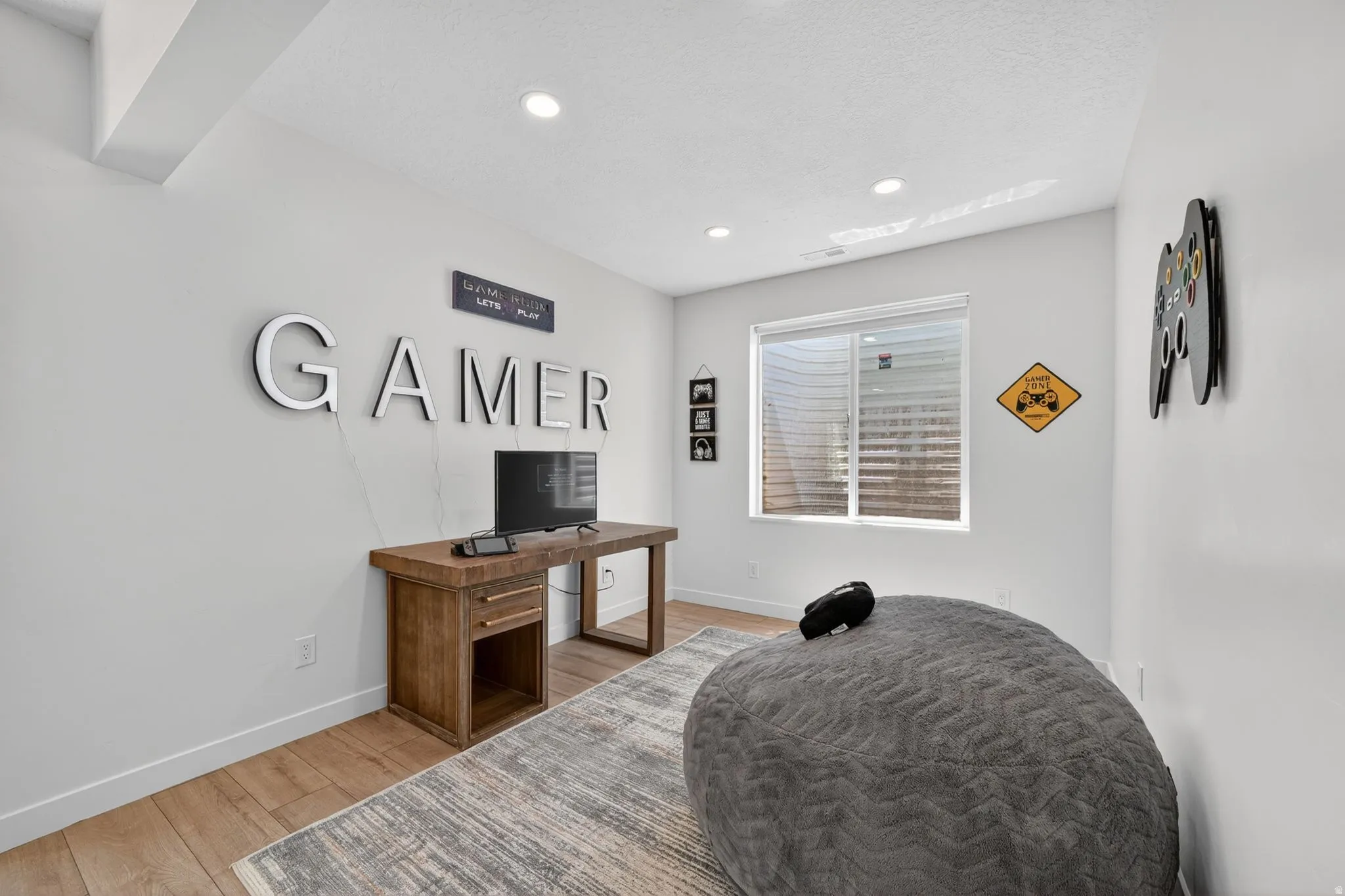 Recessed lighting illuminates this room, featuring a wood-finish desk with storage, light wood-finish flooring, and a window with blinds