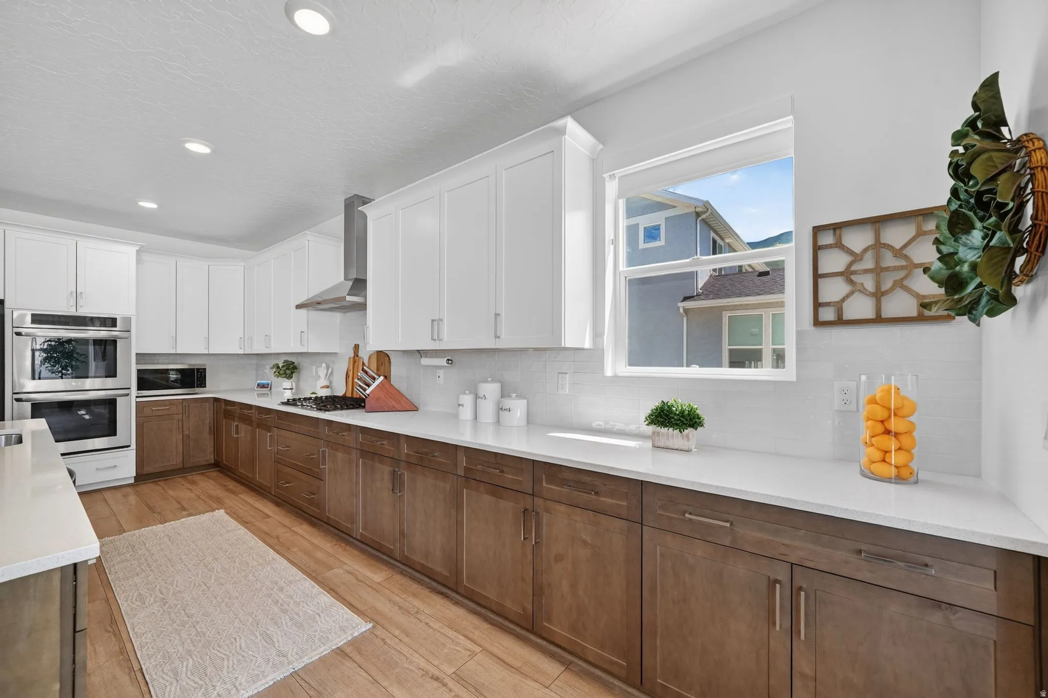 Kitchen featuring white shaker upper cabinetry and wood-finish lower cabinetry, stainless steel double wall ovens, a coordinating stainless steel range hood, and light-toned countertops