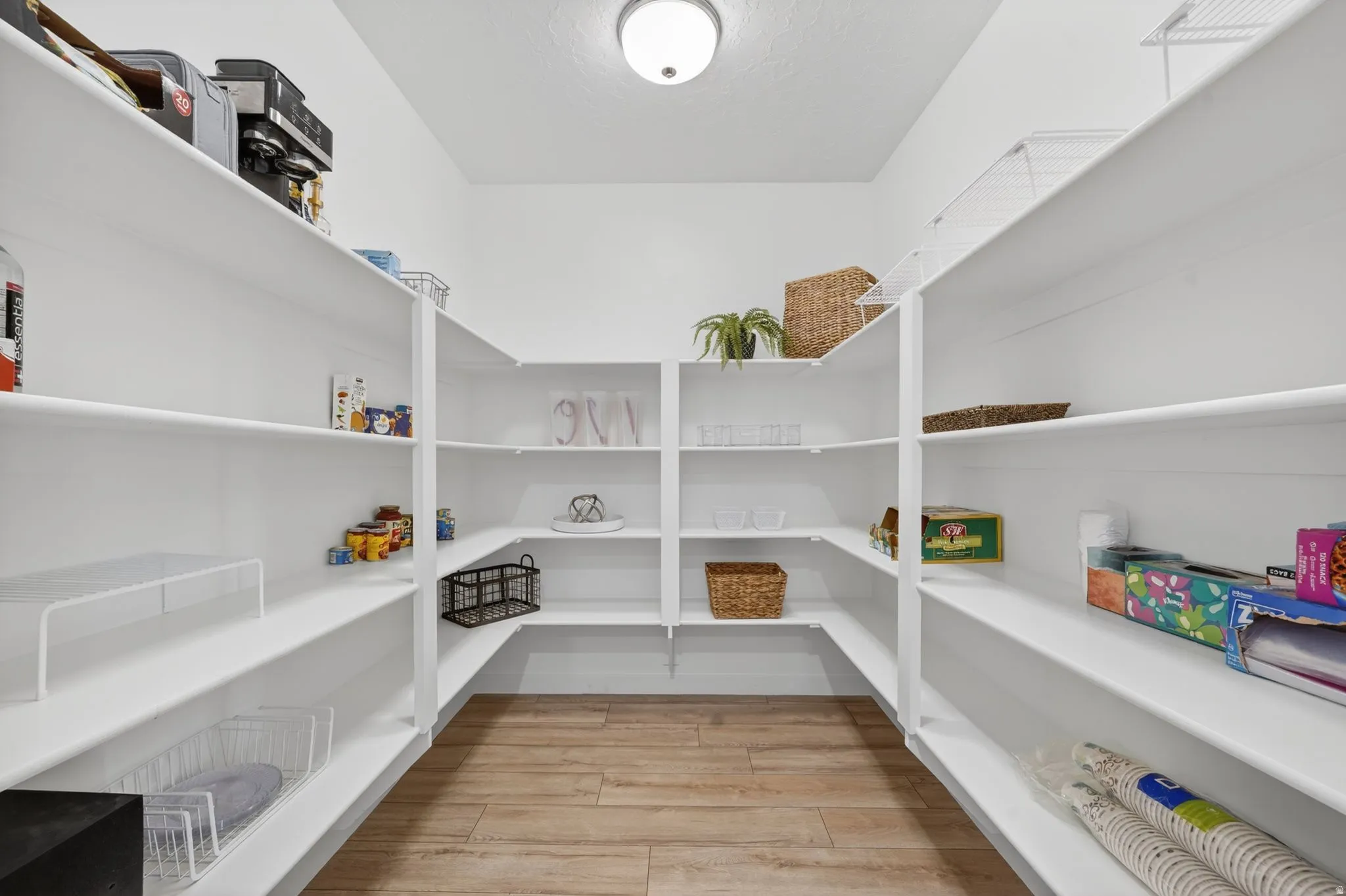 Spacious walk-in pantry featuring extensive white shelving, corner shelving units, wood-finish flooring, and a flush-mount ceiling light fixture