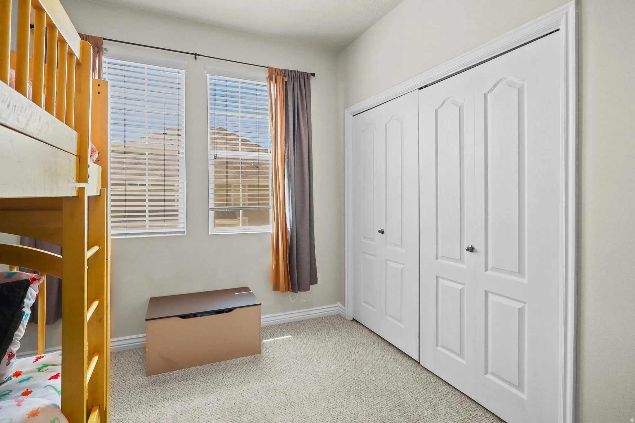 Room featuring neutral carpeting, two windows with horizontal blinds and drapery, and a bi-fold closet with panel details