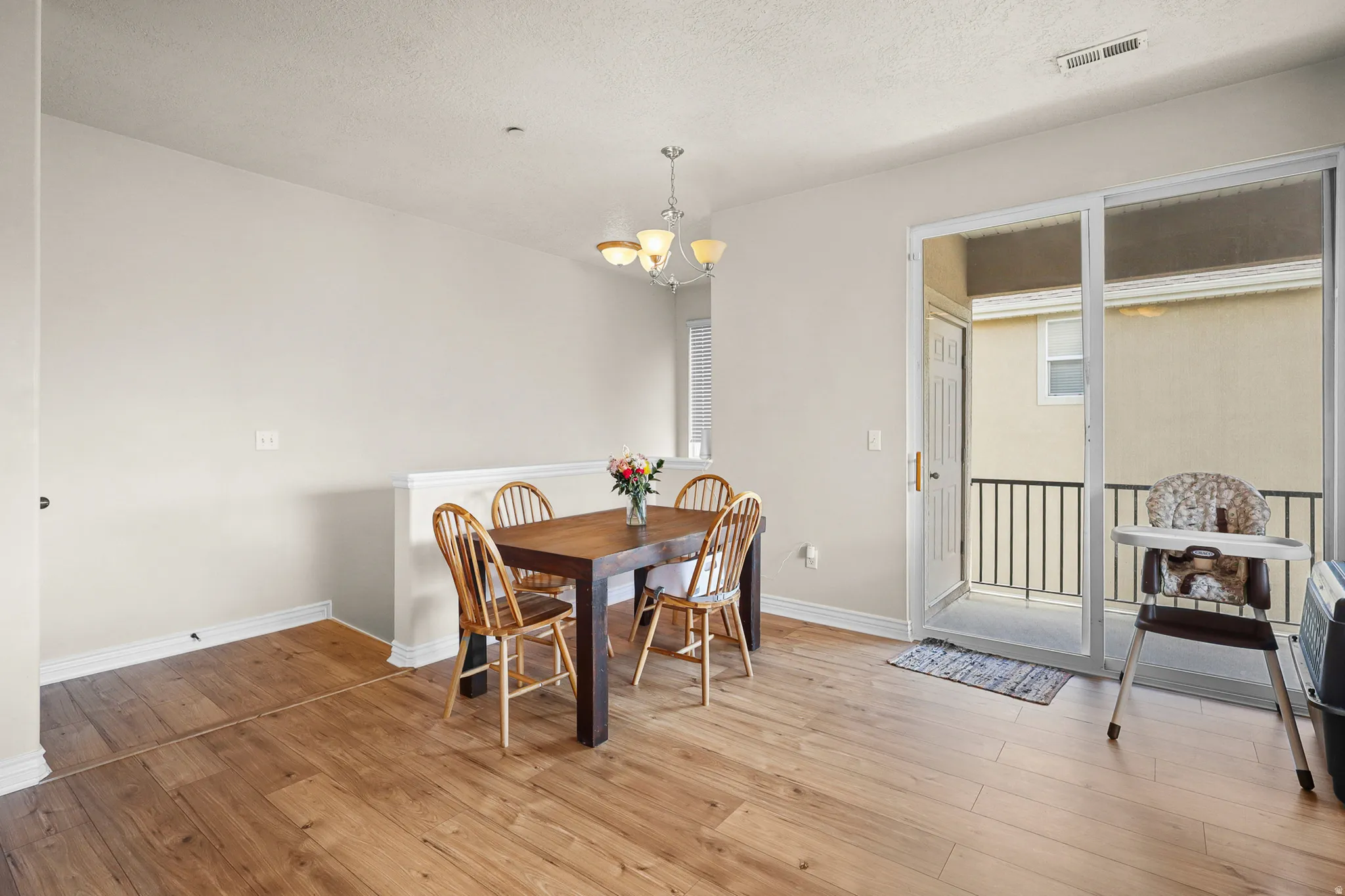 Bright interior room with wood-finish flooring, a contemporary chandelier, and a sliding glass door leading to an outdoor balcony with black metal railings