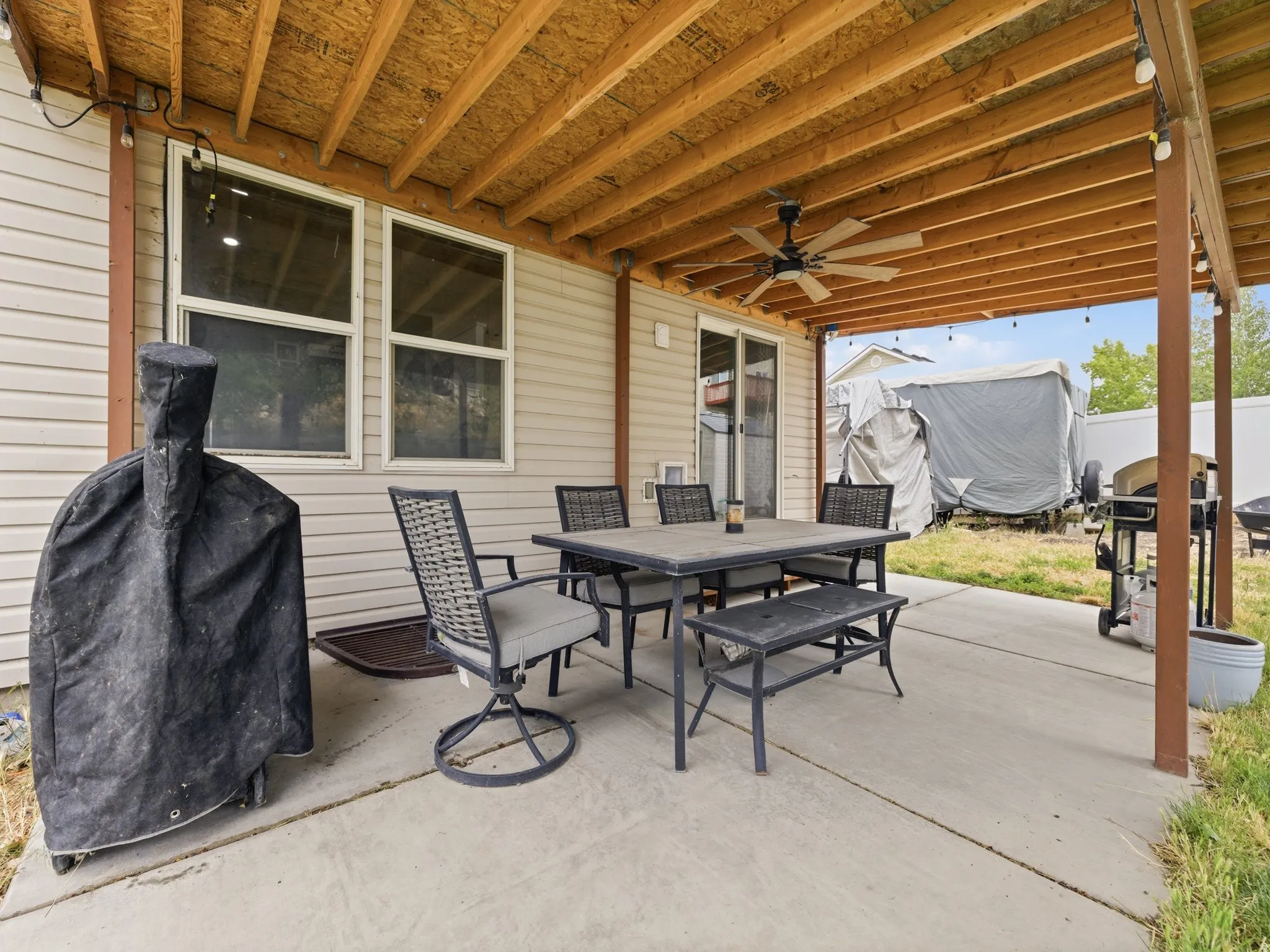 Covered concrete patio featuring an exposed wood-beam ceiling, ceiling fan, and string lighting