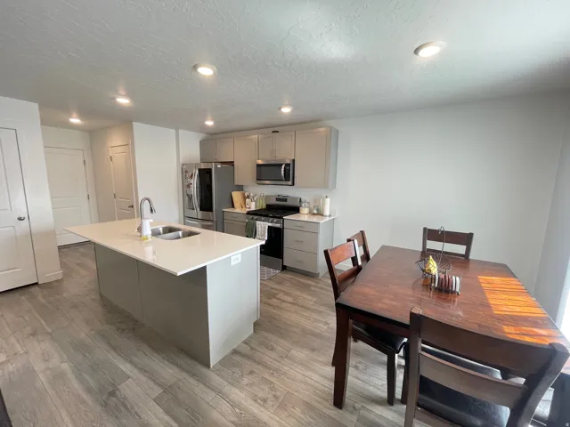 Open-concept kitchen and dining area featuring wood-finish flooring, a kitchen island with an undermount sink and gooseneck faucet, light grey cabinetry, stainless steel appliances, and recessed lighting
