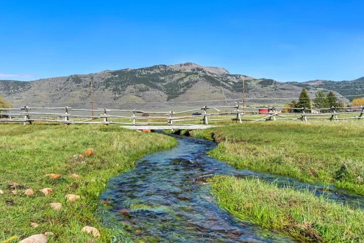 A flowing stream traversing a verdant landscape, featuring a rustic wooden fence and distant mountains under a clear sky