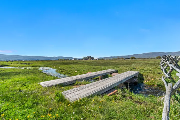 Expansive green field with a natural stream, two rustic wooden footbridges, and a split-rail fence