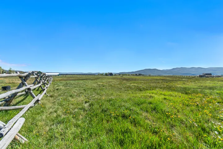 Expansive green landscape with a split-rail fence, scattered yellow wildflowers, and distant mountain ranges