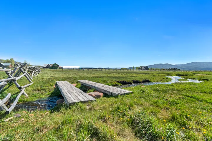 Expansive natural landscape featuring a split-rail fence, two wooden footbridges spanning a natural stream, and a distant mountain range