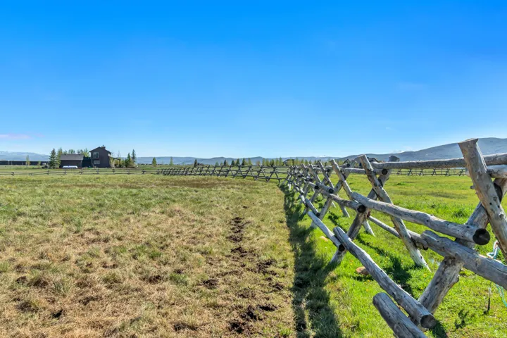 Expansive property featuring a rustic split-rail fence, verdant green grass, and a distant residence with a dark exterior