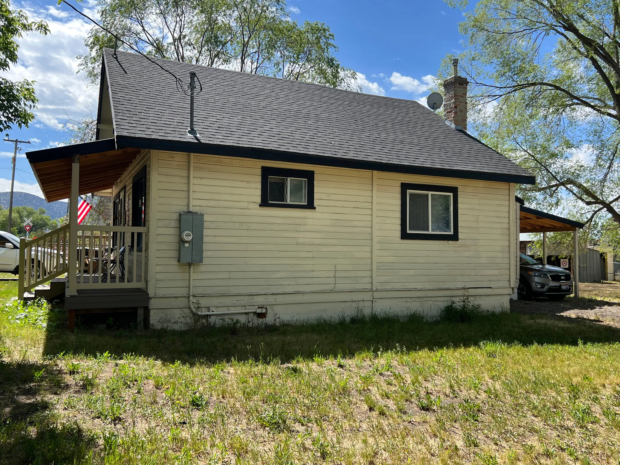 Single-story residence featuring light-colored siding, a dark shingle roof, a covered front porch with railings, a prominent brick chimney, and a covered carport area