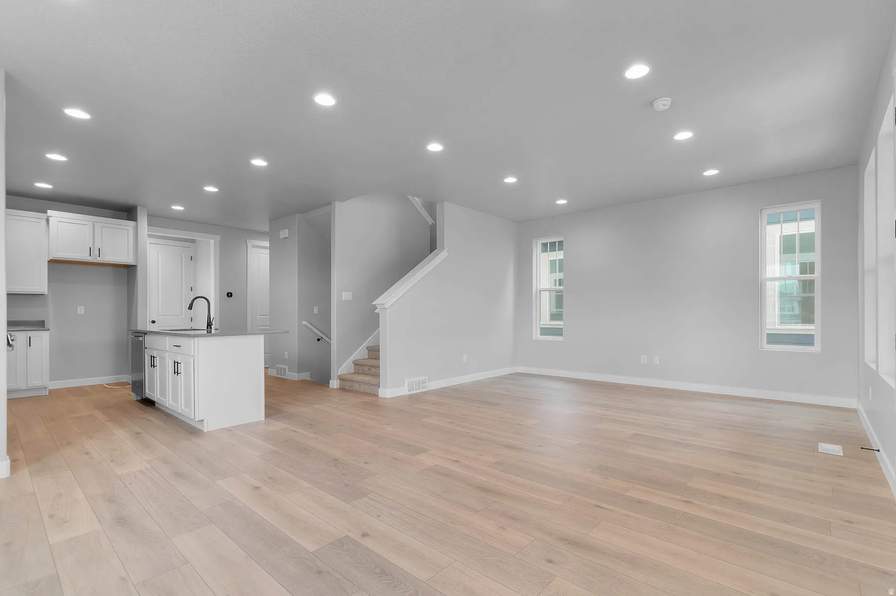 Open-concept living space featuring light wood-finish flooring, recessed lighting, white shaker cabinetry, an island with a black faucet, and multiple windows with white trim