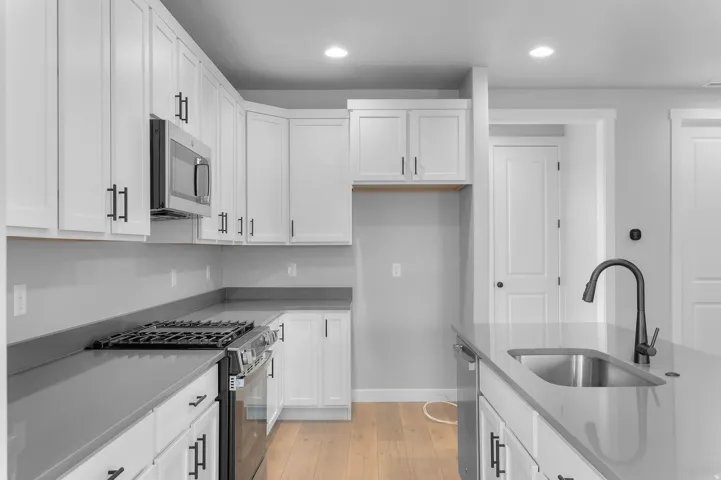 Kitchen featuring white cabinetry, dark hardware, stainless steel appliances, light wood-finish flooring, and a kitchen island with an undermount sink and matte black faucet