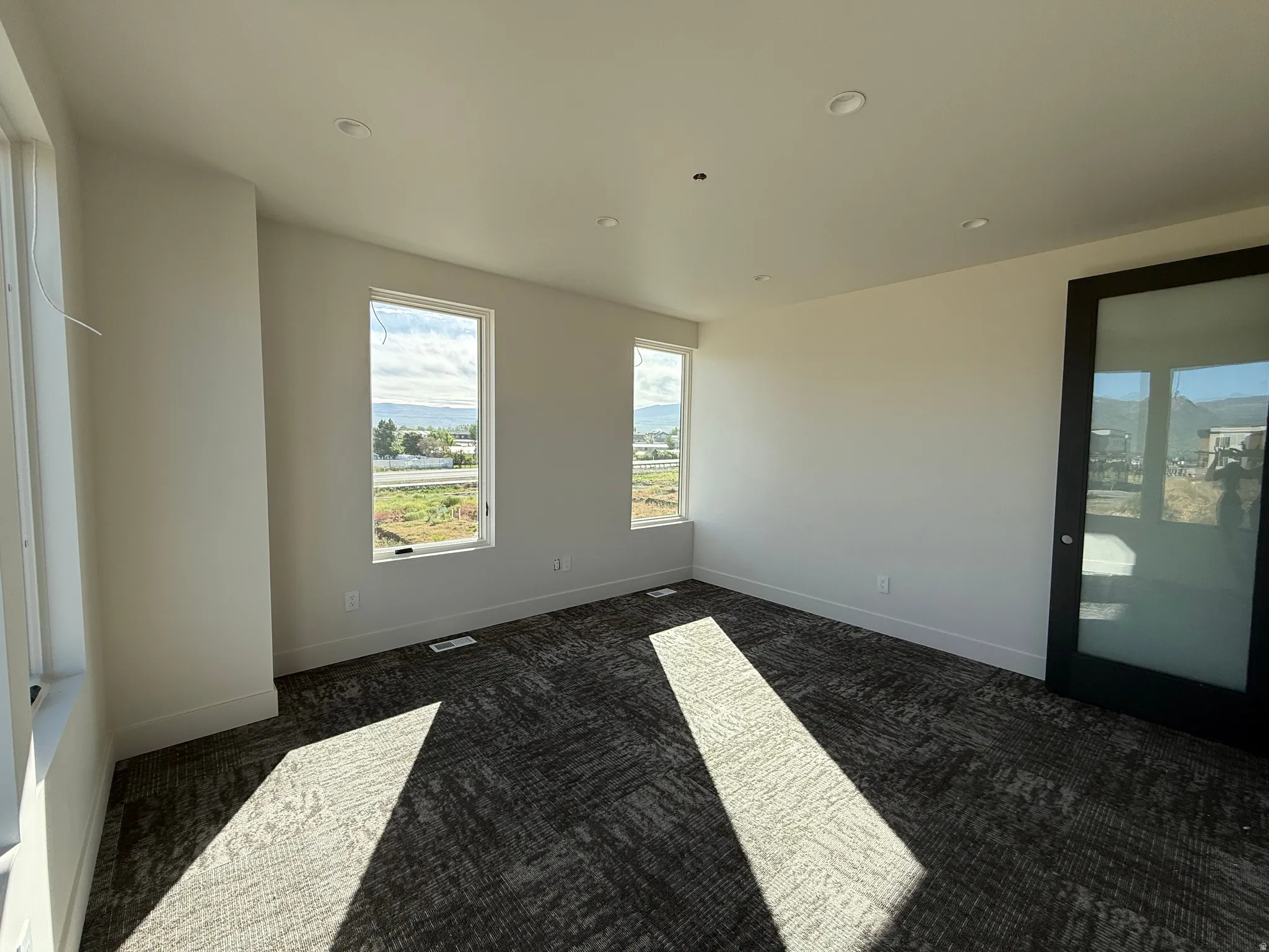 Spacious room featuring multiple large windows, recessed ceiling lighting, a modern black-framed sliding door with frosted glass, and patterned carpeting