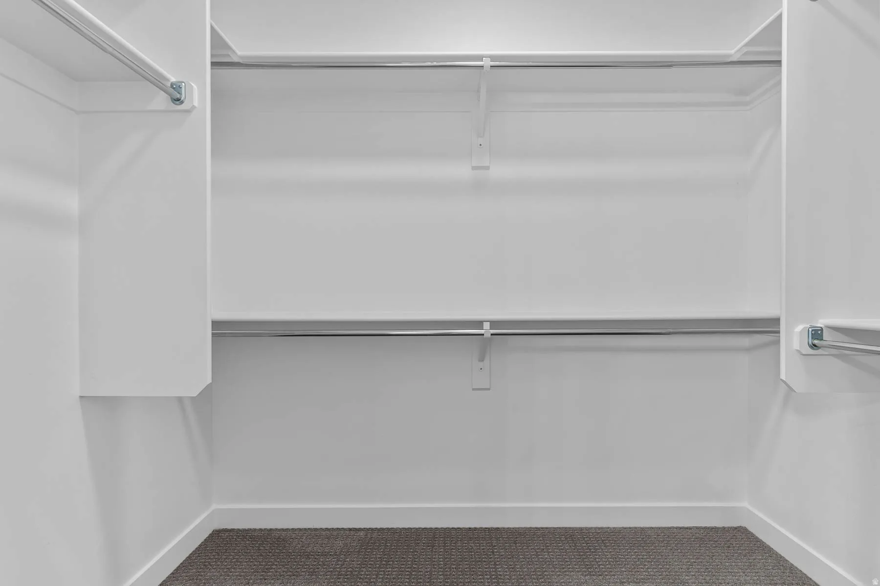 Spacious walk-in closet featuring white built-in shelving, chrome hanging rods, and dark gray carpet flooring