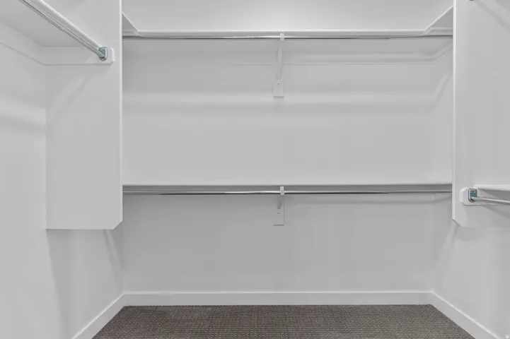 Spacious walk-in closet featuring white built-in shelving, chrome hanging rods, and dark gray carpet flooring