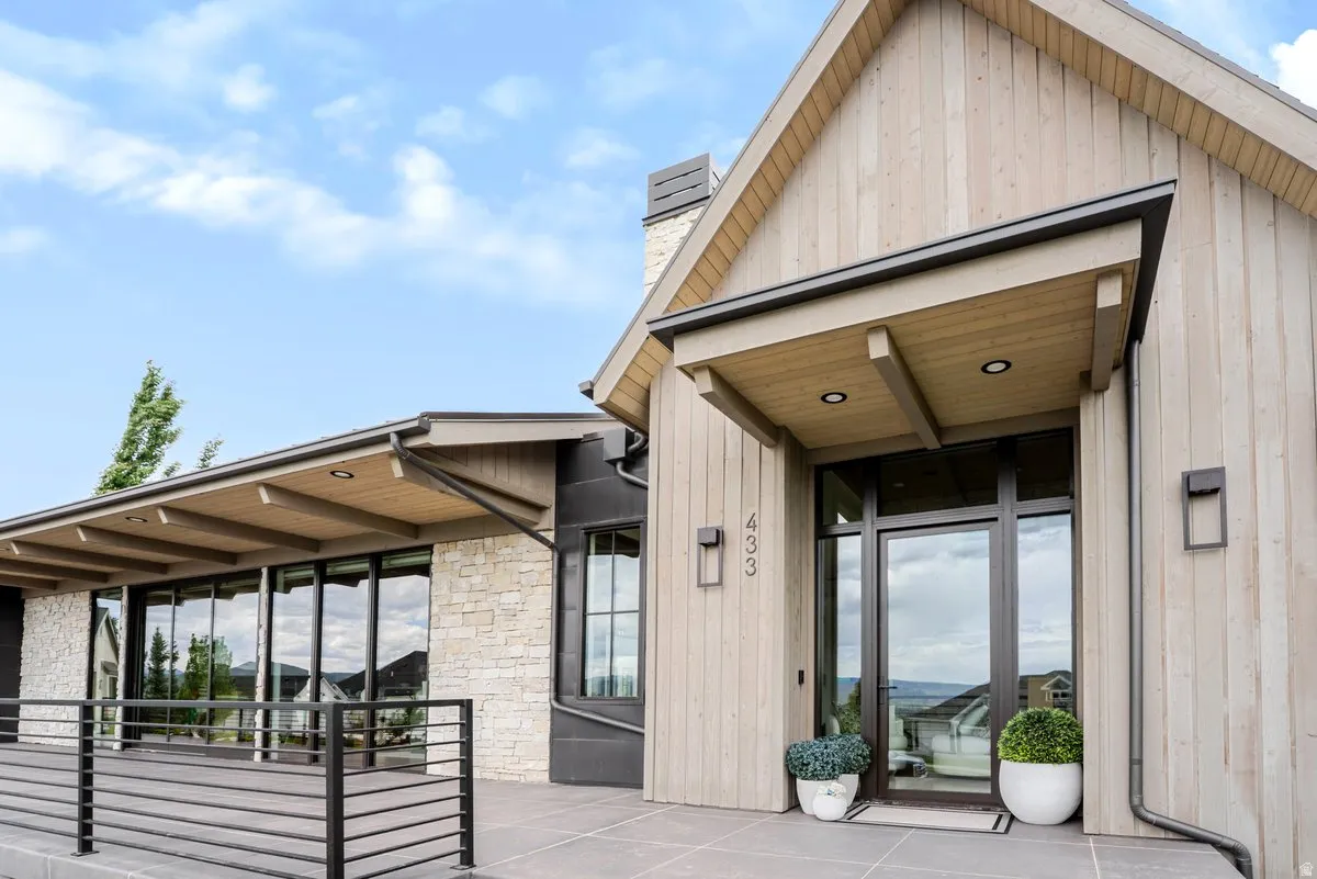 Modern architectural exterior featuring vertical wood siding, stone veneer accents, an expansive front patio with metal railing, a covered entry with recessed lighting, and large glass pane windows