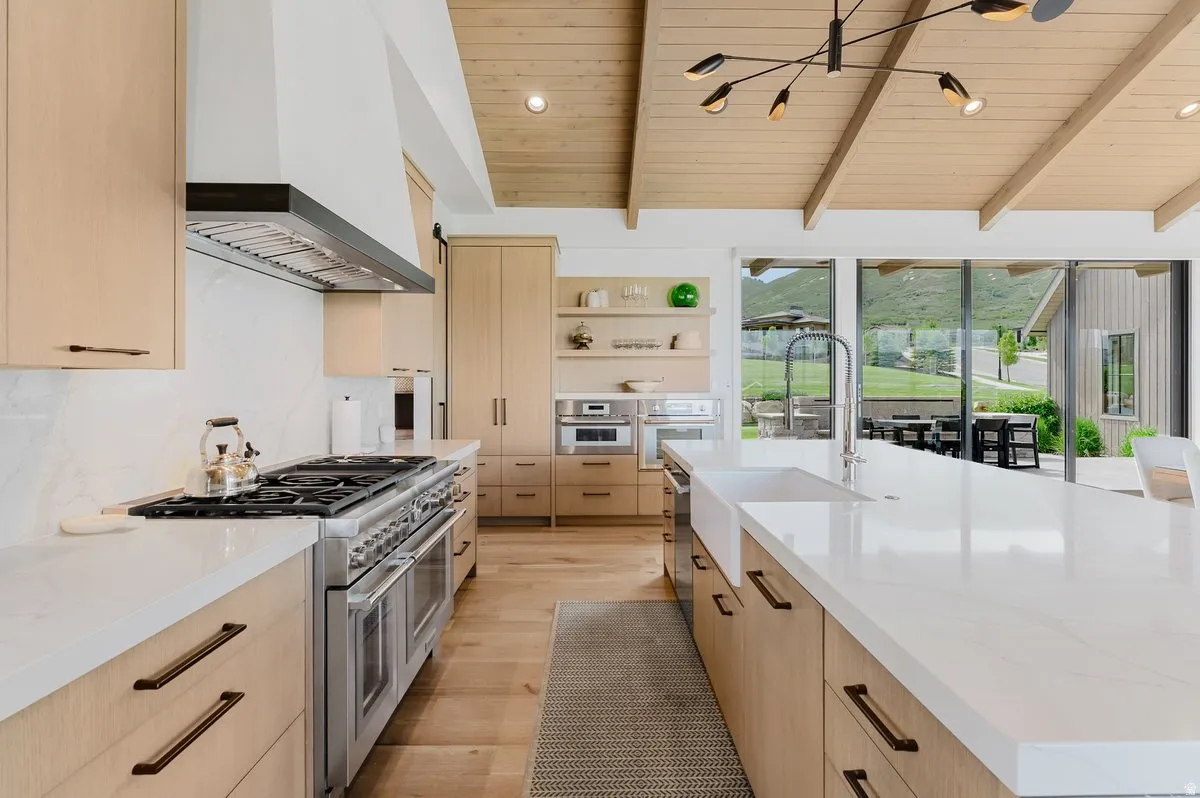 Spacious kitchen featuring a large island with a farmhouse sink, light wood-finish cabinetry, a professional-grade range with a stainless steel hood, and vaulted wood-paneled ceilings