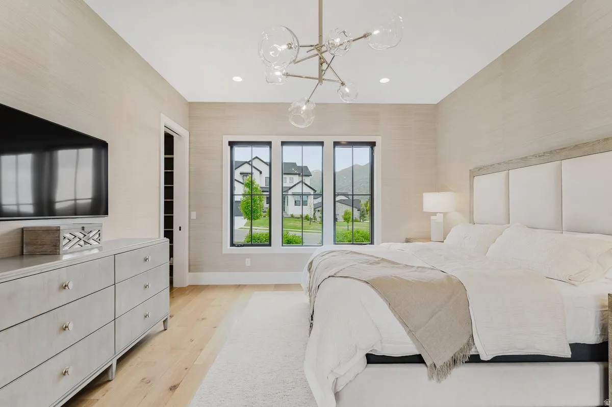 Spacious bedroom featuring light wood-finish flooring, a contemporary chandelier, a large upholstered headboard, and a three-panel window with black frames