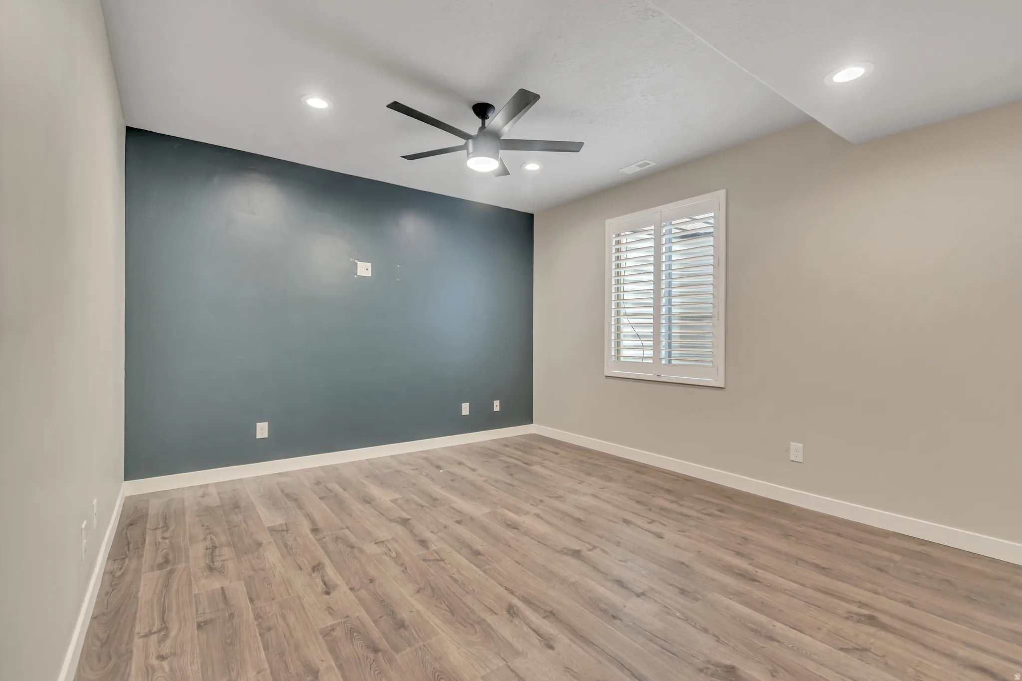 Spacious room featuring wood-finish flooring, a contemporary ceiling fan, recessed lighting, a window with white plantation shutters, and a contrasting accent wall