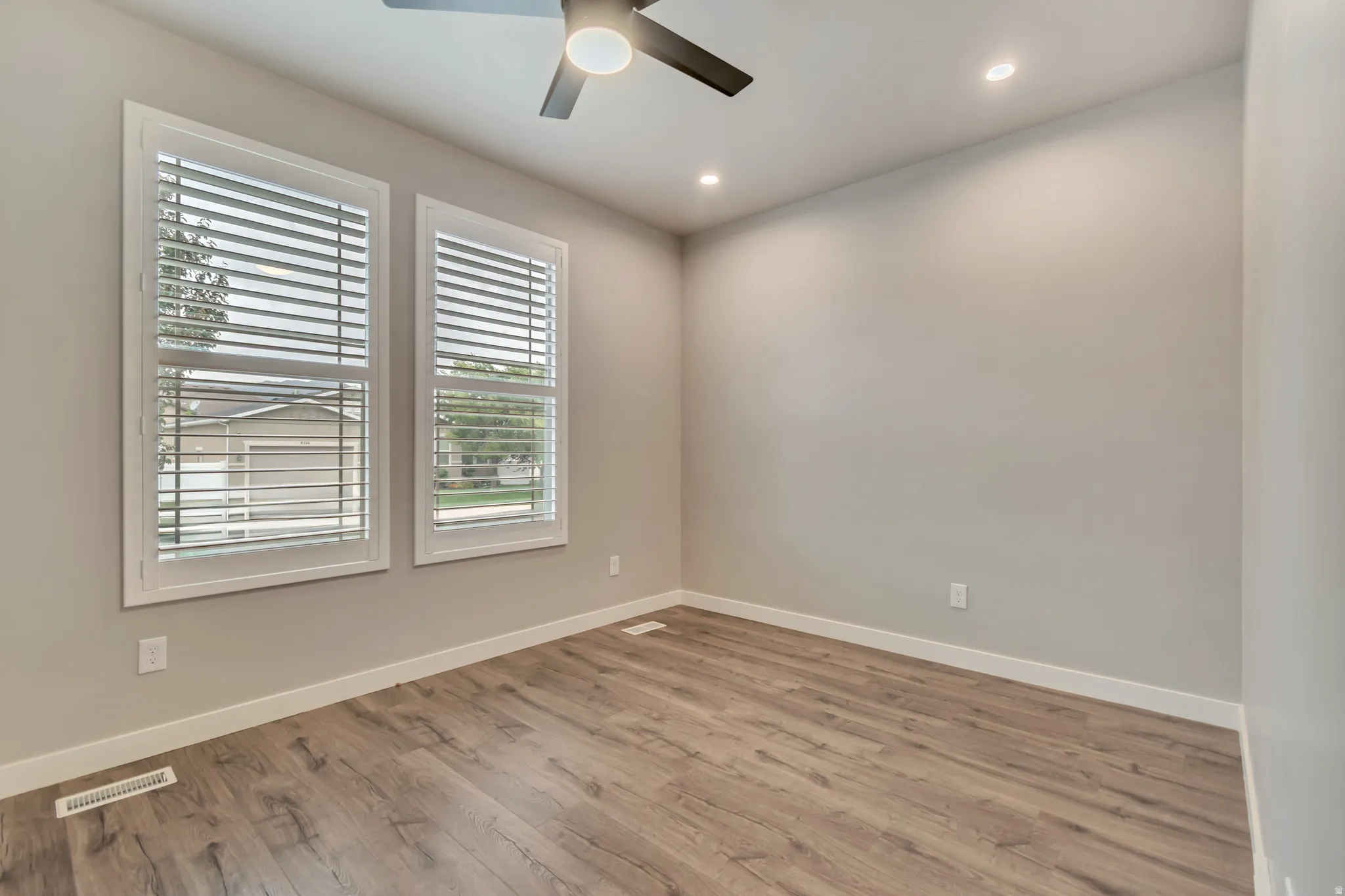 Contemporary room featuring two windows with white plantation shutters, wood-finish flooring, recessed lighting, and a ceiling fan with integrated illumination