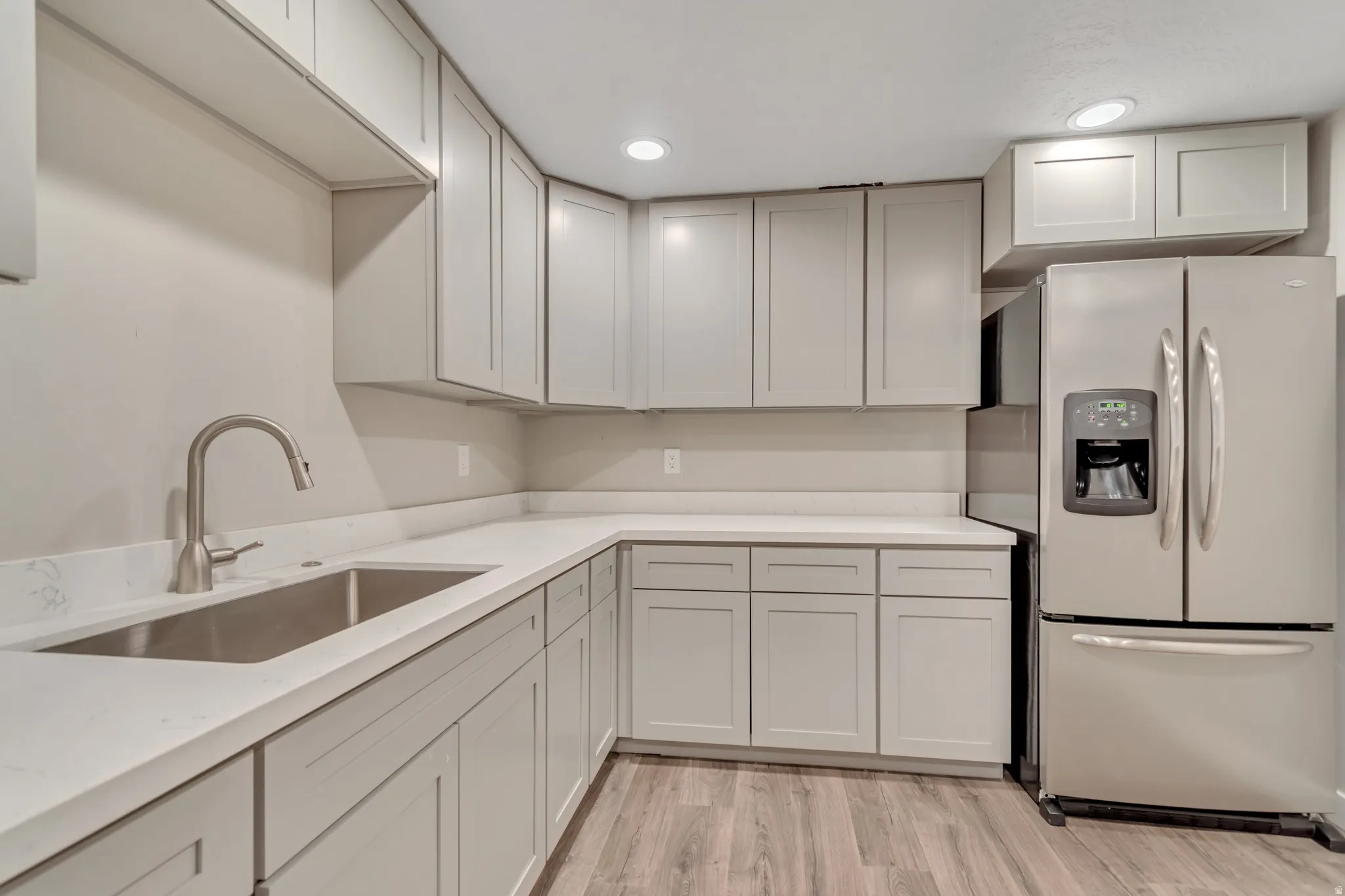Modern kitchen featuring light gray cabinetry, light-toned countertops, a stainless steel sink with a gooseneck faucet, stainless steel refrigerator, and wood-finish flooring