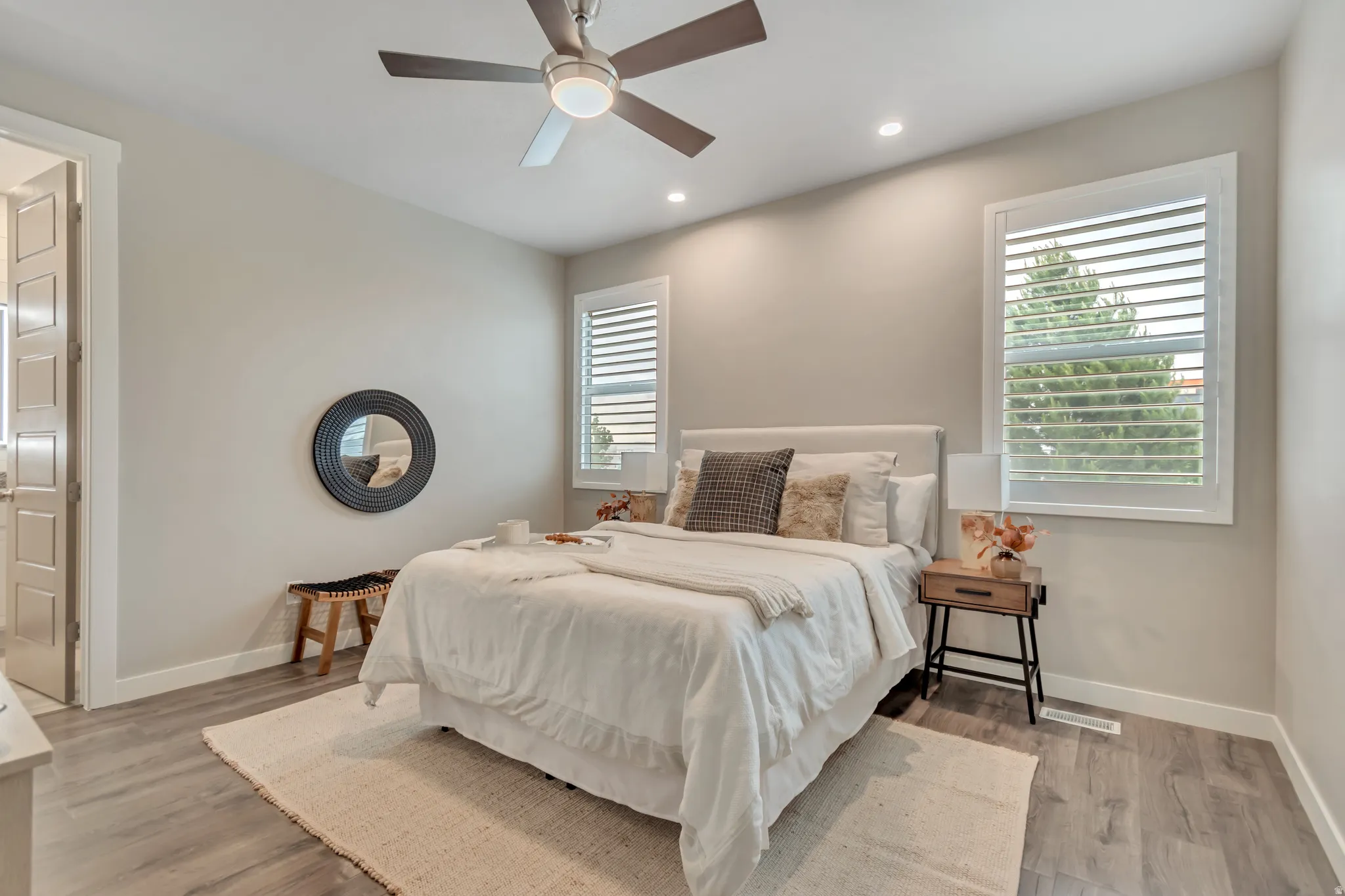 Spacious bedroom featuring wood-finish flooring, two windows with white plantation shutters, and a ceiling fan with integrated lighting