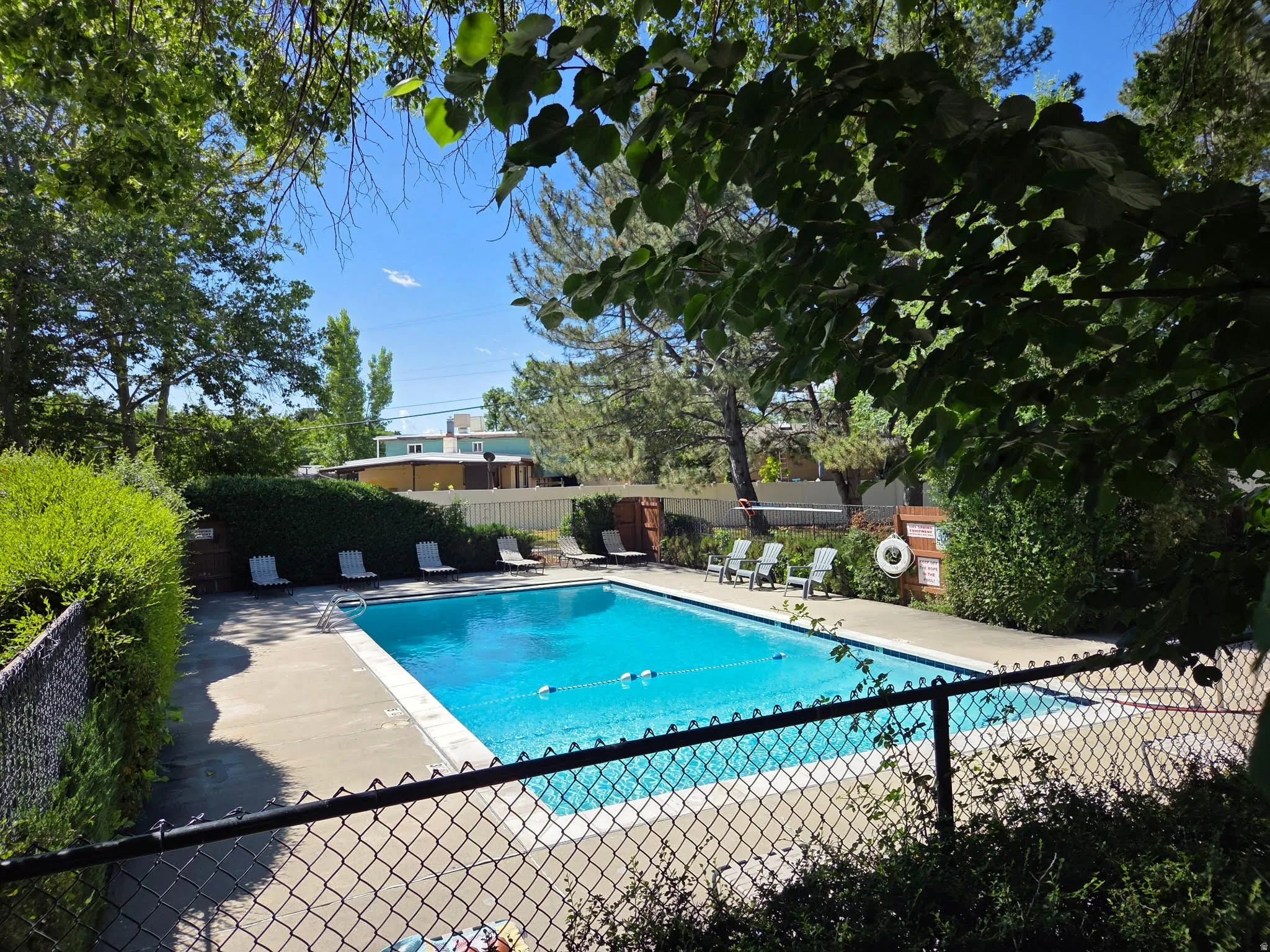 Outdoor swimming pool area featuring a perimeter fence, concrete decking, and surrounding mature landscaping