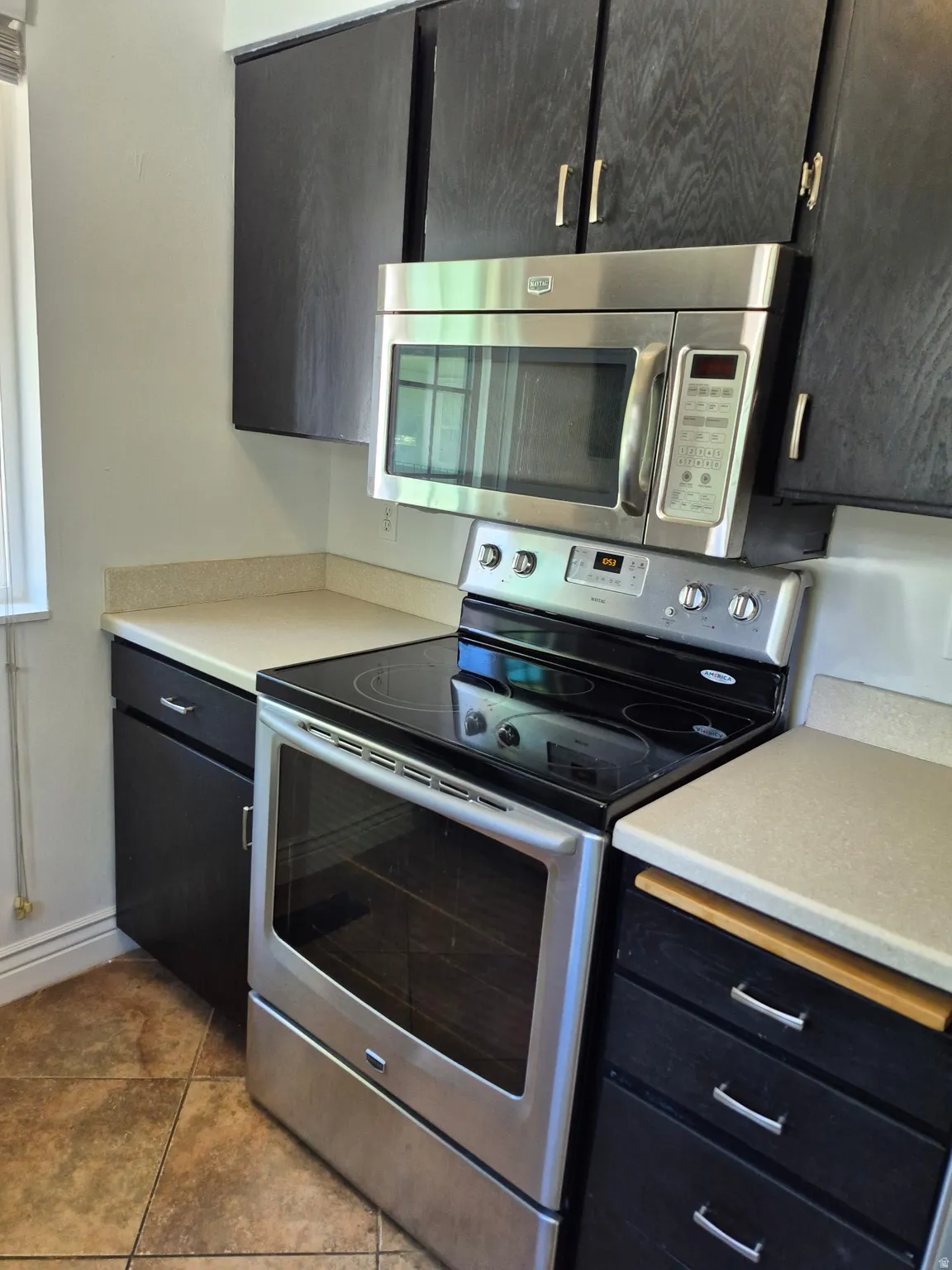 Kitchen featuring black cabinetry, stainless steel range with a smooth cooktop, coordinating stainless steel microwave, light-toned countertops, and tiled flooring