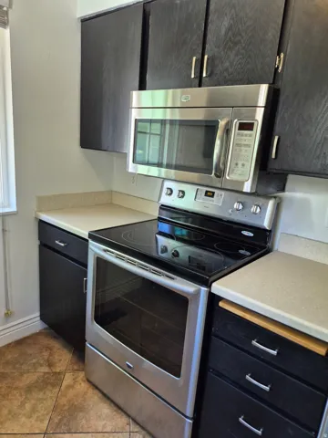 Kitchen featuring black cabinetry, stainless steel range with a smooth cooktop, coordinating stainless steel microwave, light-toned countertops, and tiled flooring