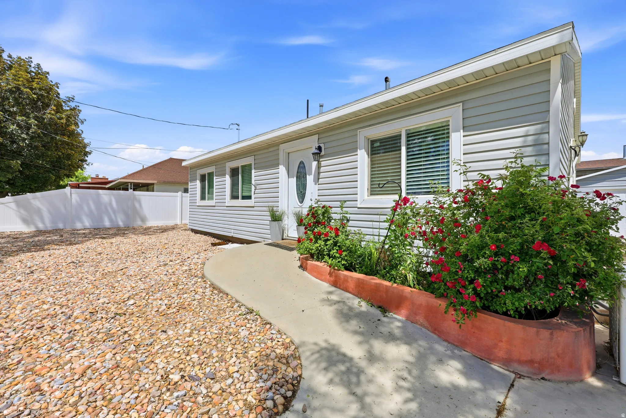 Light gray horizontal siding exterior with a white entry door featuring an oval glass insert, a concrete pathway, a low retaining wall with red flowering shrubs, and a gravel front yard area